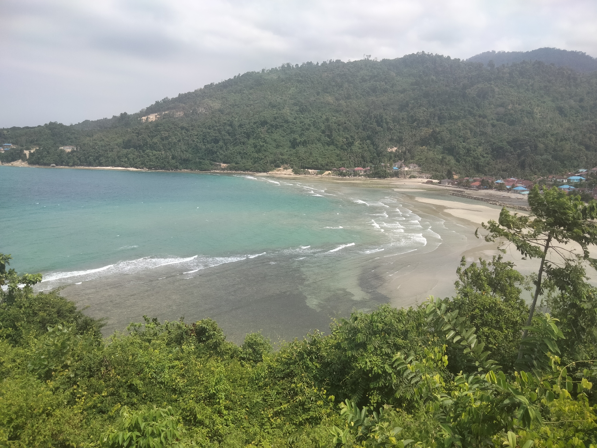 The image shows a scenic view of a tropical coastline with a lush green mountain in the background and a clear turquoise sea in the foreground. The beach is lined with a small village with colorful rooftops. The foreground is dominated by dense green foliage, suggesting a high vantage point. The sky is overcast, casting a soft, diffused light on the scene. The presence of a village suggests it is a populated area. The location is identified as Unsongi, Indonesia, a coastal town known for its natural beauty. The image captures a tranquil and idyllic setting.