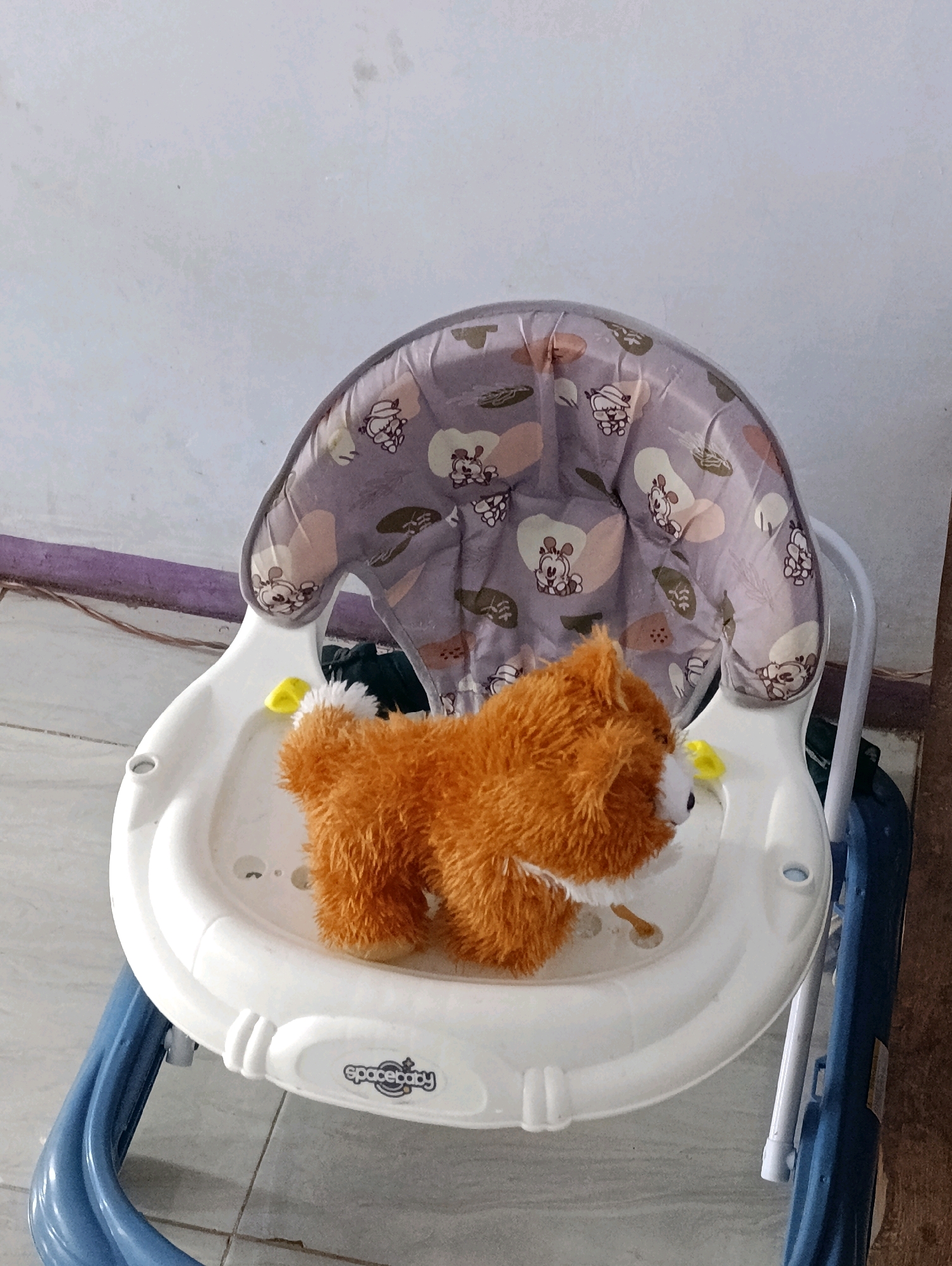 An orange plush toy dog is positioned on the white seat of a blue baby walker. The walker's seat has a grey fabric cover with a pattern of small cartoon animals and abstract shapes. A yellow toy is attached to the side of the seat. The background is a plain white wall. The scene is likely indoors, possibly in a residential setting in the city of Sebandung, Indonesia, though this cannot be definitively confirmed from the image alone.