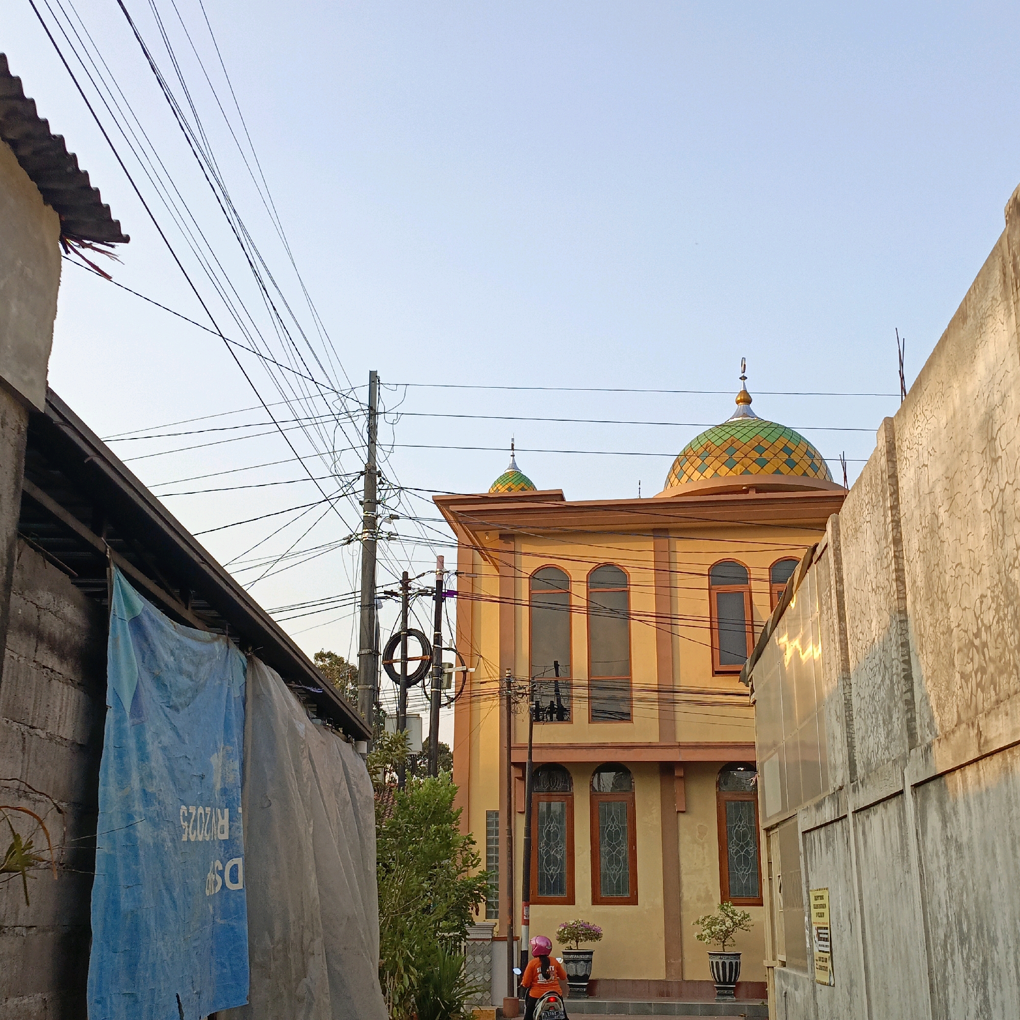 The image depicts a street scene in Salatiga, Indonesia, featuring a mosque and surrounding buildings. In the foreground, a person wearing an orange shirt and pink helmet rides a motorcycle, viewed from behind. To the left, a section of a building is covered by blue and grey tarpaulins, with visible brickwork. A utility pole with a complex network of overhead wires is prominent on the left side, extending towards the center. The central focus is a yellow mosque with a distinctive green and yellow tiled dome, featuring arched windows with decorative grills. To the right, a textured concrete wall, possibly a boundary wall or building facade, runs along the street. The sky is clear and pale blue, suggesting it might be late afternoon due to the warm light reflecting on the right-side wall.