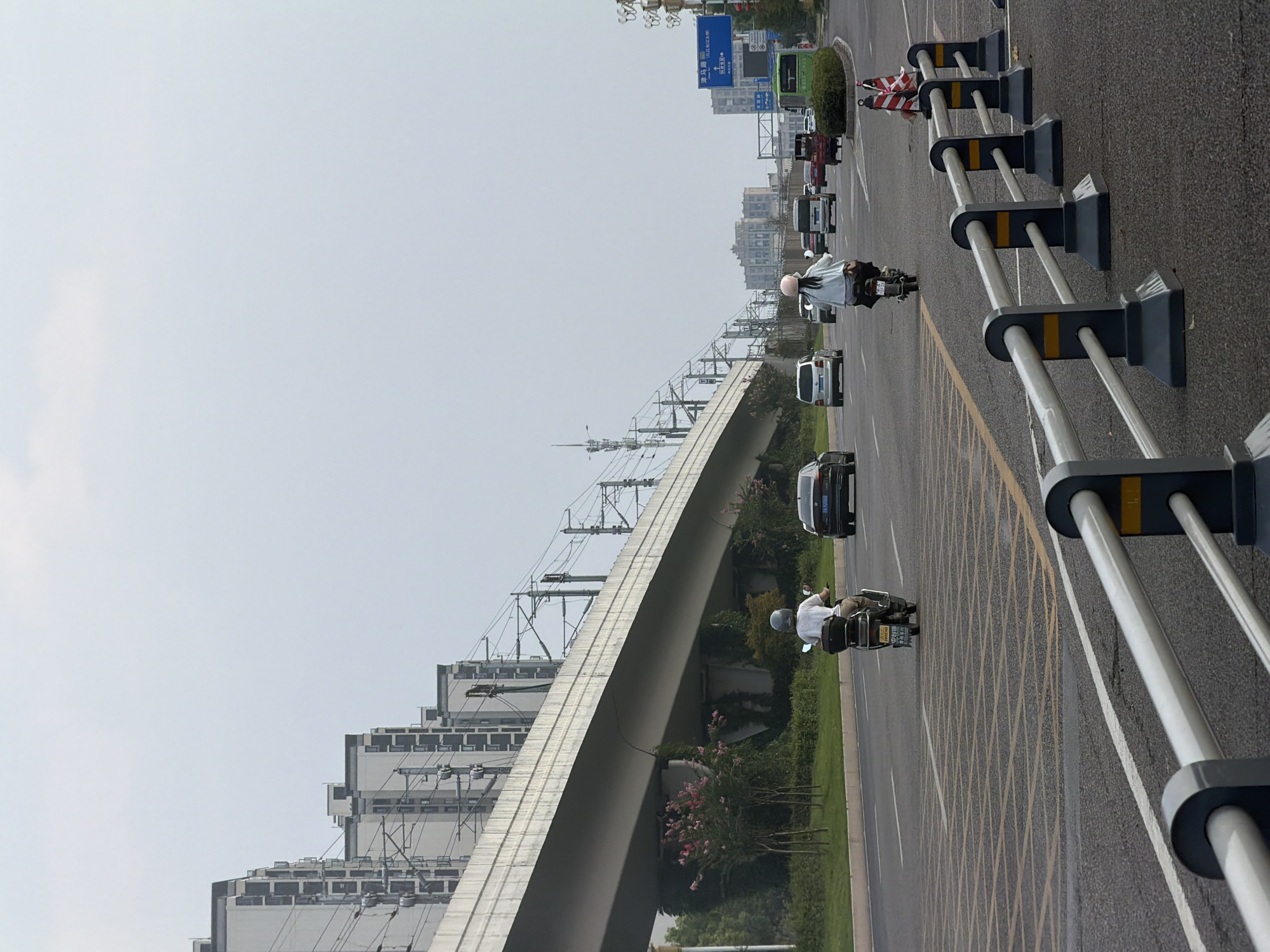 The image shows a city scene in Jiangjin, China, with an elevated structure resembling a highway or train track in the background. Several residential buildings are visible under the structure. In the foreground, a road with multiple lanes is depicted. A person is riding a scooter in the right lane, facing away from the camera. Another person on a scooter is in the left lane, also facing away. Several cars and a green bus are on the road. Traffic barriers with orange and white stripes are placed along the side of the road. The sky is overcast.