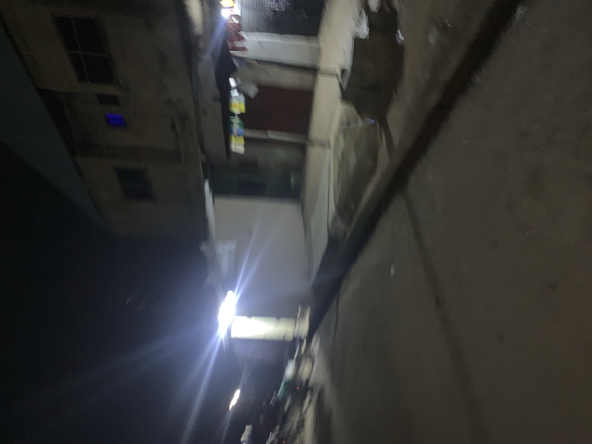 The image depicts a night scene on a street in Gura Top, Nigeria. A bright light source, likely a streetlamp, illuminates the area from the left, casting strong lens flares. In the background, there is a two-story building with several windows. One window on the upper floor emits a distinct blue light. Below the upper floor, a covered walkway or balcony area is visible, with clothes hanging on a line. Red doors are visible on the ground floor of the building. The street itself appears to be paved, with a darker drainage channel running along the side. Out of focus on the left side of the image, there are indications of what appear to be motorcycles or bicycles. The overall scene is slightly blurred, consistent with a low-light, potentially handheld, photograph.