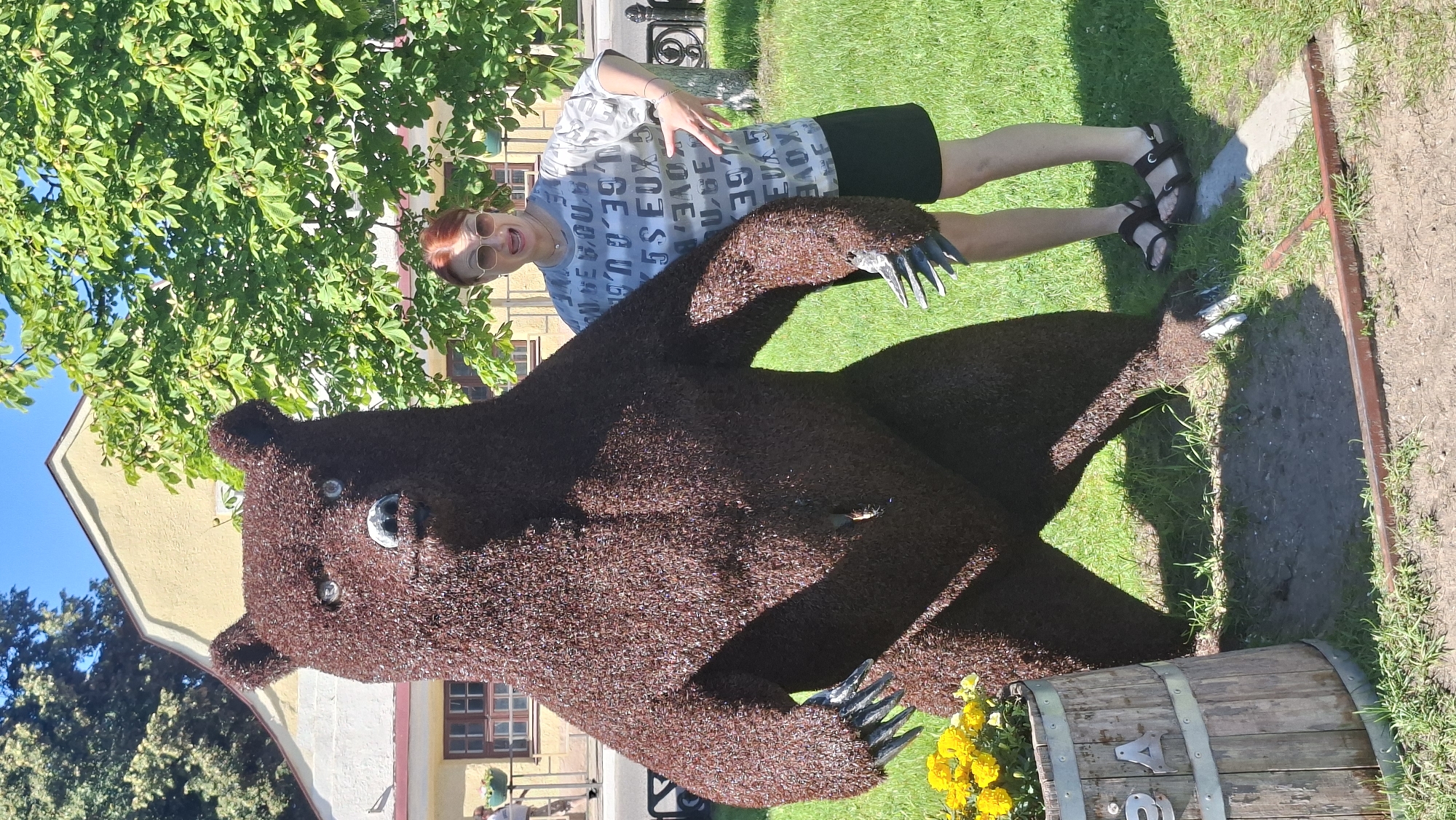 A woman with red hair and sunglasses stands to the right of a large, brown, wire sculpture of a bear. The woman is wearing a grey t-shirt with a repeating pattern of letters and a black skirt. She is smiling and has her right hand raised with fingers spread. The bear sculpture is standing upright with its arms down. To the left of the bear sculpture, a wooden barrel filled with yellow flowers is visible. In the background, there is a yellow building with windows and a tree with green leaves. The scene is outdoors on a sunny day, with green grass covering the ground. The location is the city of Pastavy, Belarus, as indicated by text on the barrel, which appears to say "IPA".