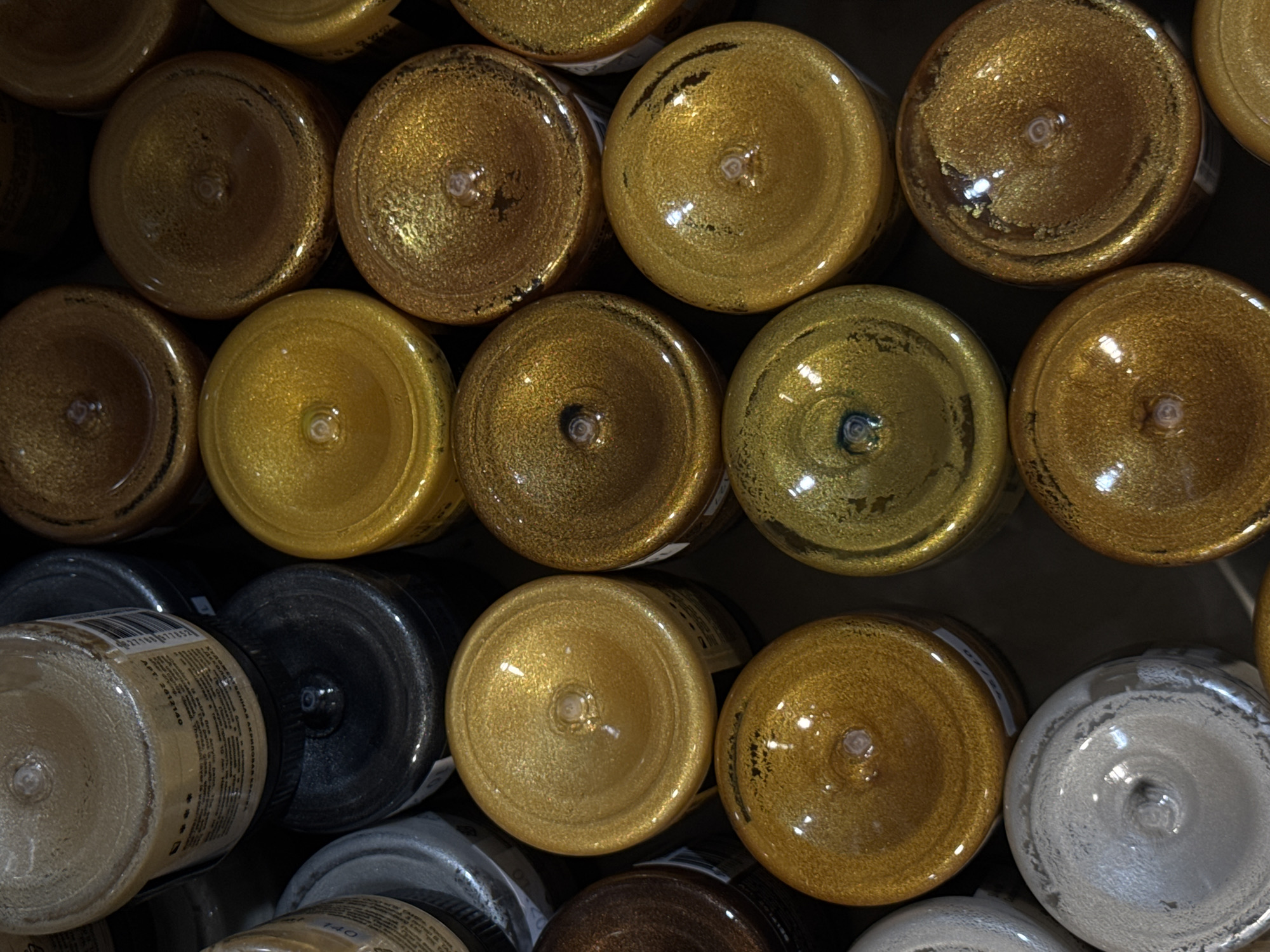 This media file displays an overhead, close-up view of numerous bottles, primarily filled with metallic gold paint, tightly packed together. The bottles are of various sizes and shapes, but most appear to be small to medium-sized containers with screw-on lids, typical for craft or art supplies. The contents of the bottles are shimmering and reflective, suggesting they contain glitter or metallic pigments. The arrangement of the bottles is dense, with some bottles partially obscuring others, creating a visually textured pattern. The lighting appears to be artificial, likely from an indoor setting, as there are distinct highlights and reflections on the glossy surfaces of the bottles and their contents.
The scene is one of organized storage, possibly in a retail environment, a craft studio, or a workshop. There are no people visible in the frame. The focus is entirely on the objects themselves. The variety of gold tones, from bright yellow-gold to deeper, antique gold, suggests a range of product options. In the lower left corner, one bottle is partially visible with a label that has printed text, including what appears to be a barcode and some Cyrillic characters, consistent with the stated location context of Moscow, Russia. The overall impression is one of abundant artistic or crafting materials.