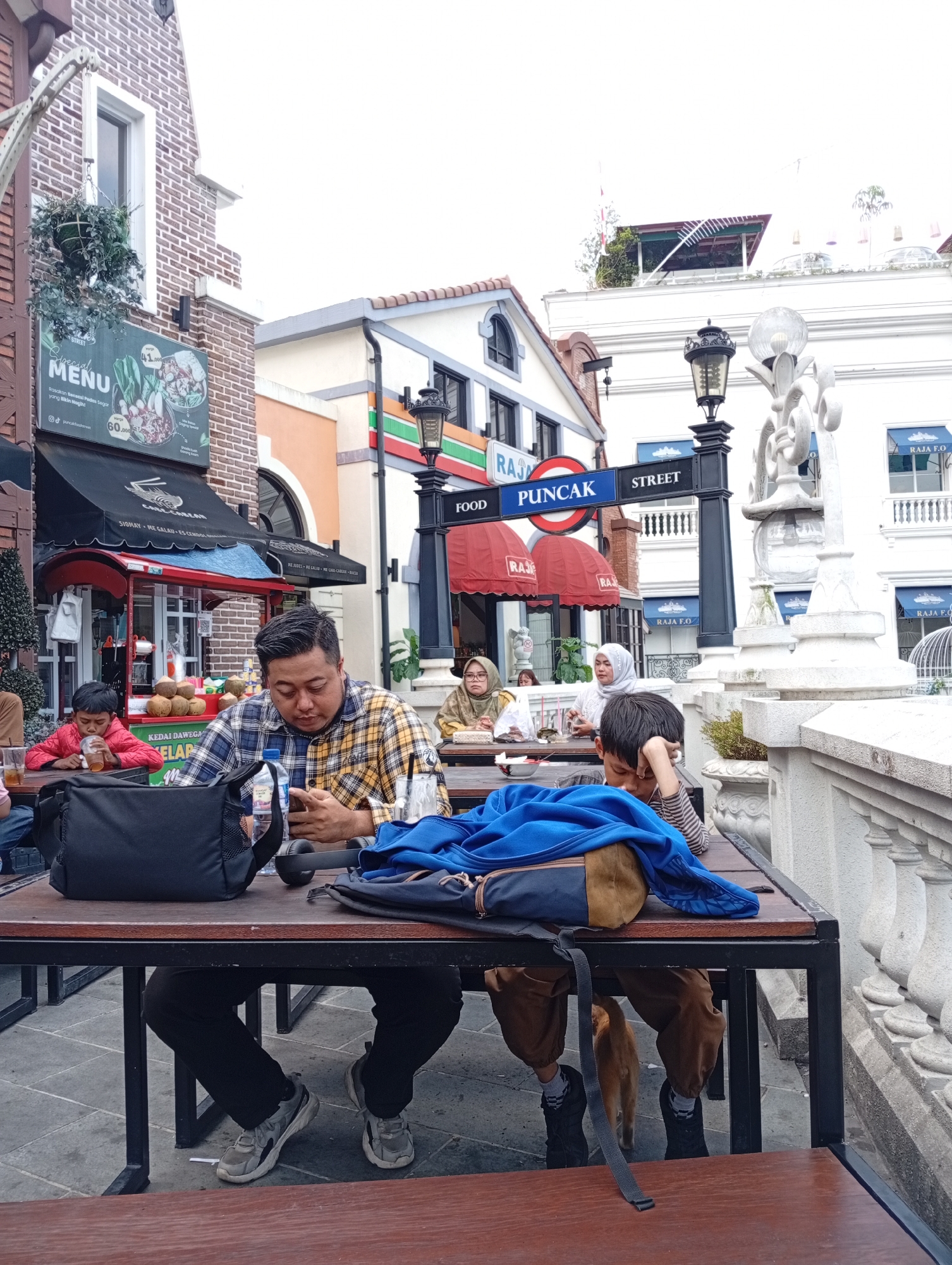 The image depicts an outdoor dining area, likely part of a commercial establishment in Cimacan, Indonesia. In the foreground, a man in a plaid shirt is seated at a table, looking down at his phone. To his right, a child in a red jacket is also seated at the table, holding a drink. Another child, partially obscured by a blue blanket on the table, is also present. In the background, several other individuals are dining at tables, and the architecture suggests a European-inspired theme with brick buildings and decorative street lamps. Signs indicate "FOOD" and "STREET" in English, as well as a "MENU" sign in Indonesian. The overall impression is that of a casual dining experience in a developed tourist area.