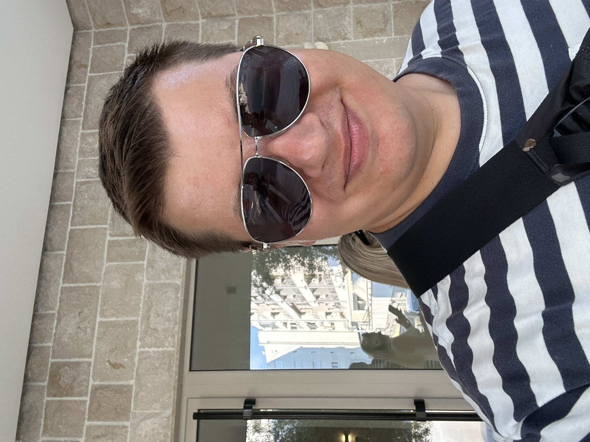 The media file is a portrait of a man wearing sunglasses and a striped shirt. He is outdoors, with a stone-tiled wall in the background. A window reflects buildings and trees, suggesting an urban or suburban setting. The man's face is the primary focus, with his sunglasses obscuring his eyes. His lips are slightly parted. He is wearing a dark and white striped t-shirt, and a black strap crosses his chest. The image appears to have been taken during the daytime, with natural light illuminating the scene. The reflection in the window shows a sunny day. The overall mood is casual.