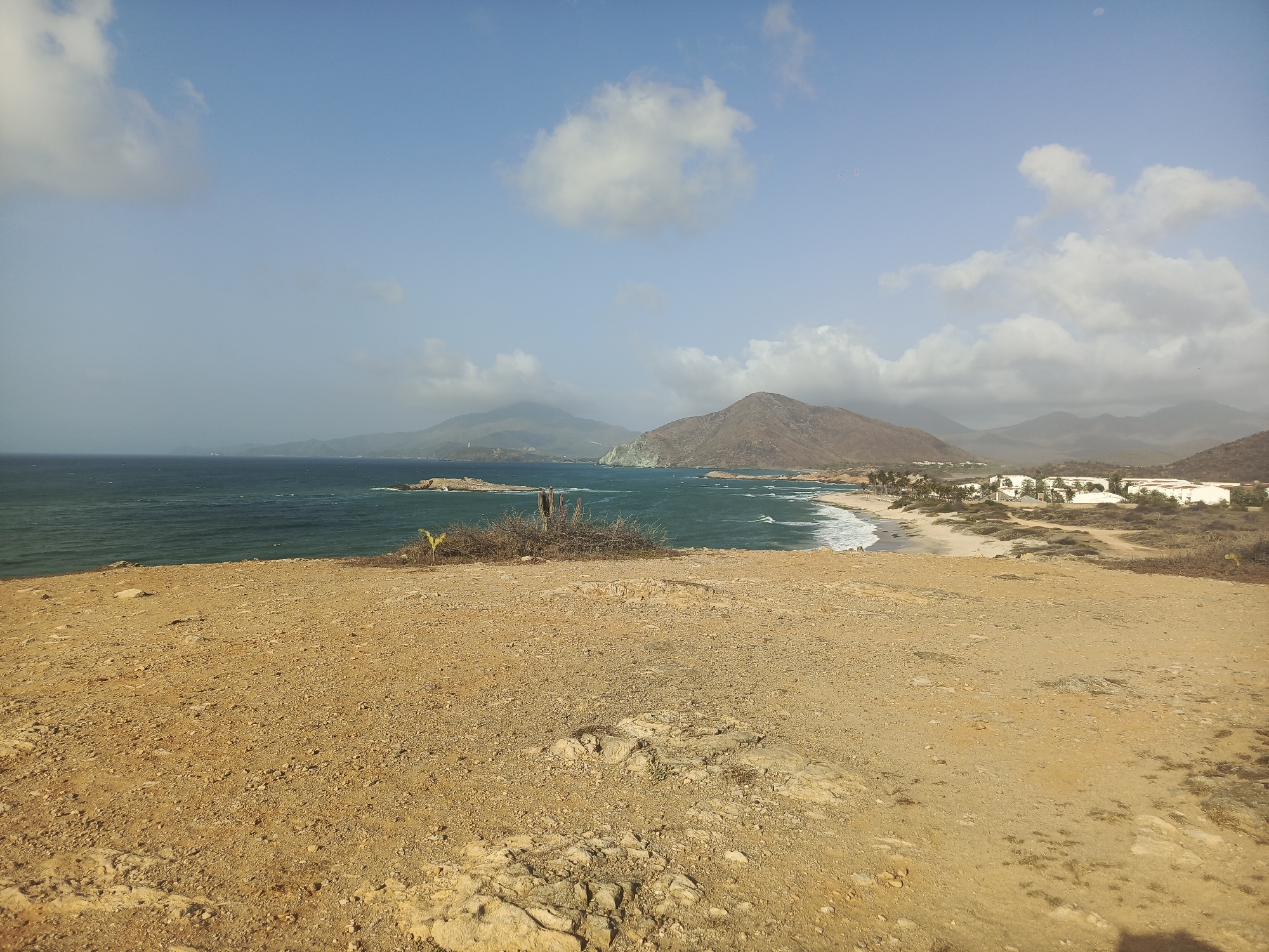 The image presents a landscape in Altagracia, Venezuela. The foreground consists of a dry, rocky terrain with sparse vegetation. In the middle ground, a coastline with a sandy beach is visible, bordered by the turquoise water of the Caribbean Sea. The background features hills and mountains under a blue sky with scattered clouds. Some structures resembling houses or buildings are visible along the beach. There are no people visible in the image. The overall impression is of a coastal scene in a dry, mountainous region.