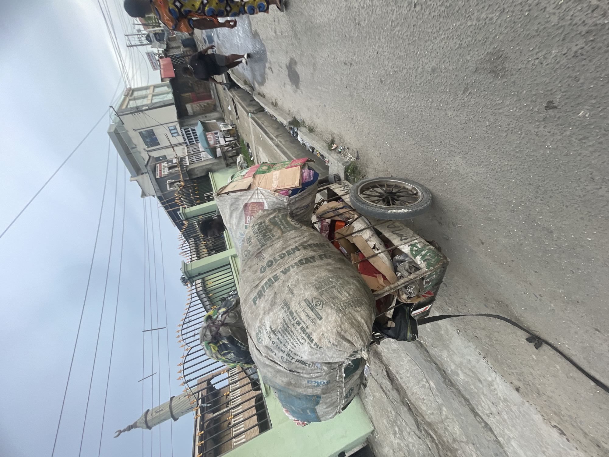 An outdoor street scene in Ado, Nigeria, is captured on what appears to be an overcast day. The image, presented as if rotated 90 degrees counter-clockwise from its original orientation, primarily features a heavily laden, makeshift cart or trolley on the right side of a paved road.
The cart is filled with various items, including cardboard boxes and other refuse, suggesting it's used for collecting materials, possibly for recycling or scrap. Dominating the cart's load is a large, grey woven sack prominently displaying text. Visible text on the sack includes "PRIME WHEAT FLOUR," "GOLDEN," and manufacturing details such as "FLOUR MILLS OF NIGERIA PLC," "Old Dock Road, Apapa, Lagos, Nigeria," and "www.fmplc.com." Other discernible phrases on the sack are "MFO DATE," "BEST BEFORE DATE," and instructions to "Store in a cool dry place." A single wheel of the cart is visible on the right.
To the left of the cart, the paved road extends into the background, where a drainage ditch or gutter filled with some debris runs parallel to the street. In the mid-ground, a person wearing dark clothing walks away from the viewer. Another individual, partially visible in the upper background, wears brightly patterned attire.
Buildings of varying styles line the left side of the street. One structure has visible railings or balconies, while another in the distance features a distinct finial or decorative element, possibly from a mosque's minaret. Overhead, numerous power lines or utility cables crisscross against the bright, grey sky. A small sign with the word "HERE" is visible in the background, near one of the buildings. A black strap or cable lies on the ground next to the cart. The overall scene depicts a glimpse into daily life in an urban or semi-urban environment.