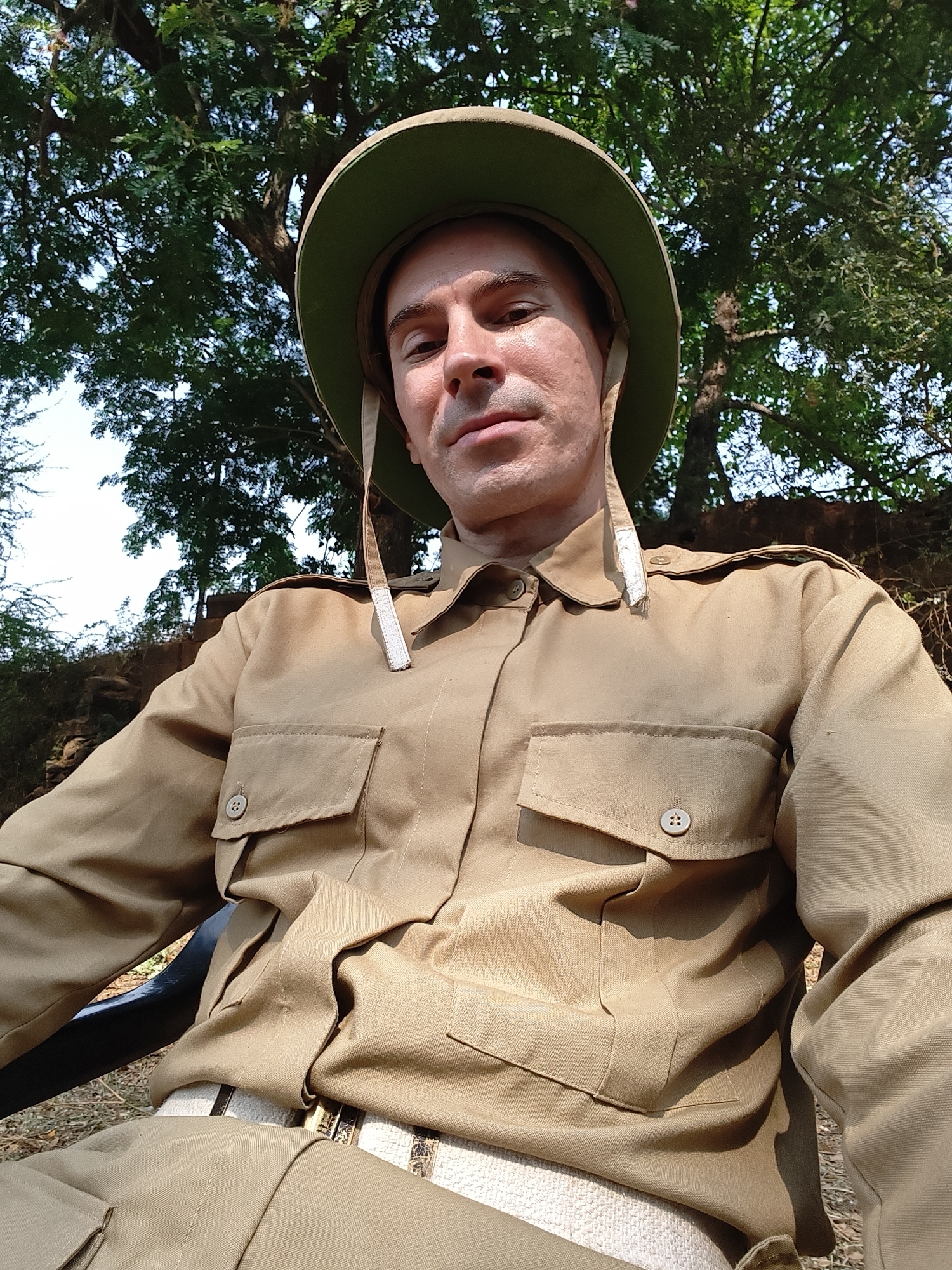 An adult Caucasian male is depicted in a seated selfie taken outdoors in Kolhapur, India. He wears a light olive-green pith helmet with a beige brim underside and chin straps, a tan/khaki long-sleeved collared shirt with two buttoned breast pockets and shoulder epaulets, and matching tan/khaki trousers. A distinct white woven belt with a gold-toned buckle is visible at his waist. He looks directly at the camera with a neutral expression. A black armrest of a chair is visible supporting his left arm. The background features dense green tree foliage against a bright sky, with a stone or earthen embankment covered in dry vegetation visible on the mid-right. The ground appears dry with sparse debris.
