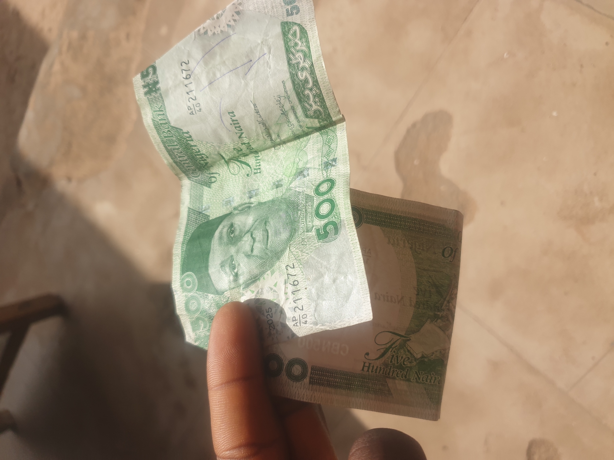 The image shows a close-up of a Nigerian 500 Naira note being held by a person's hand. The note is green and features a portrait of Nnamdi Azikiwe. Some of the text visible on the note includes "Central Bank of Nigeria," "Five Hundred Naira," and the serial number "AP 40 211672." The note is creased and slightly worn. The background appears to be a textured, sandy surface, possibly a ground or floor, with some dappled sunlight suggesting an outdoor setting. The image is taken from a slightly high angle, looking down at the note and the hand. There is no indication of the city of Jauro Yinu, Nigeria in the image.