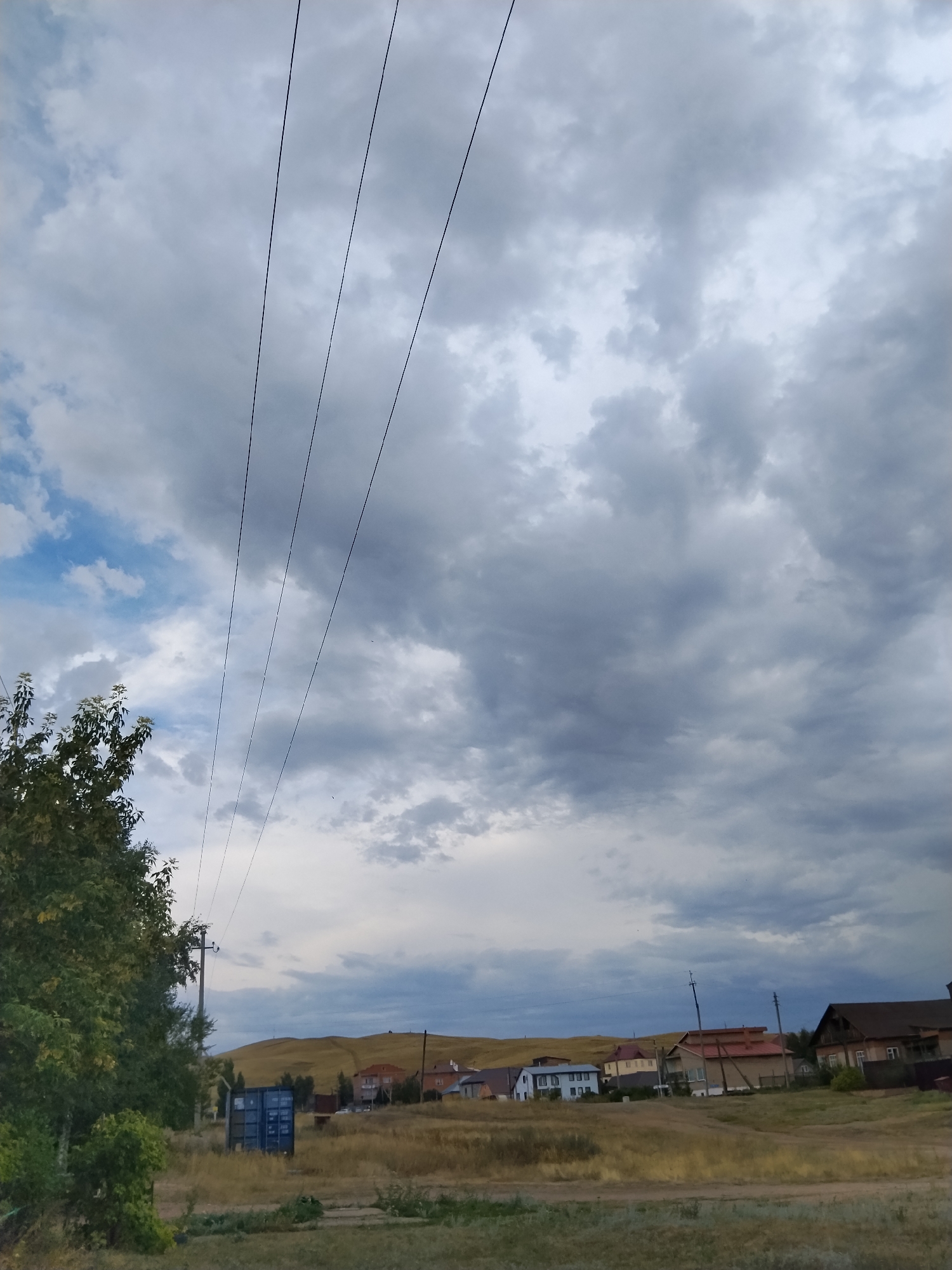 The image depicts a rural landscape under a cloudy sky, likely on the outskirts of Kokshetau, Kazakhstan. In the foreground, a grassy area with some scattered dry vegetation borders a dirt path. To the left, a dense green tree with some lighter foliage is partially visible. A bright blue shipping container stands in the middle ground. Further back, a collection of houses with various roof colors, including red and brown, are nestled on a gentle, grassy slope that leads up to a distant, rounded hill. Utility poles with power lines are visible in the scene, with the wires stretching diagonally across the upper portion of the frame. The sky is filled with a mixture of light and dark gray clouds, with some patches of blue visible on the left side. No people or animals are present in the image. The overall impression is one of a quiet, semi-developed rural area under an overcast sky.