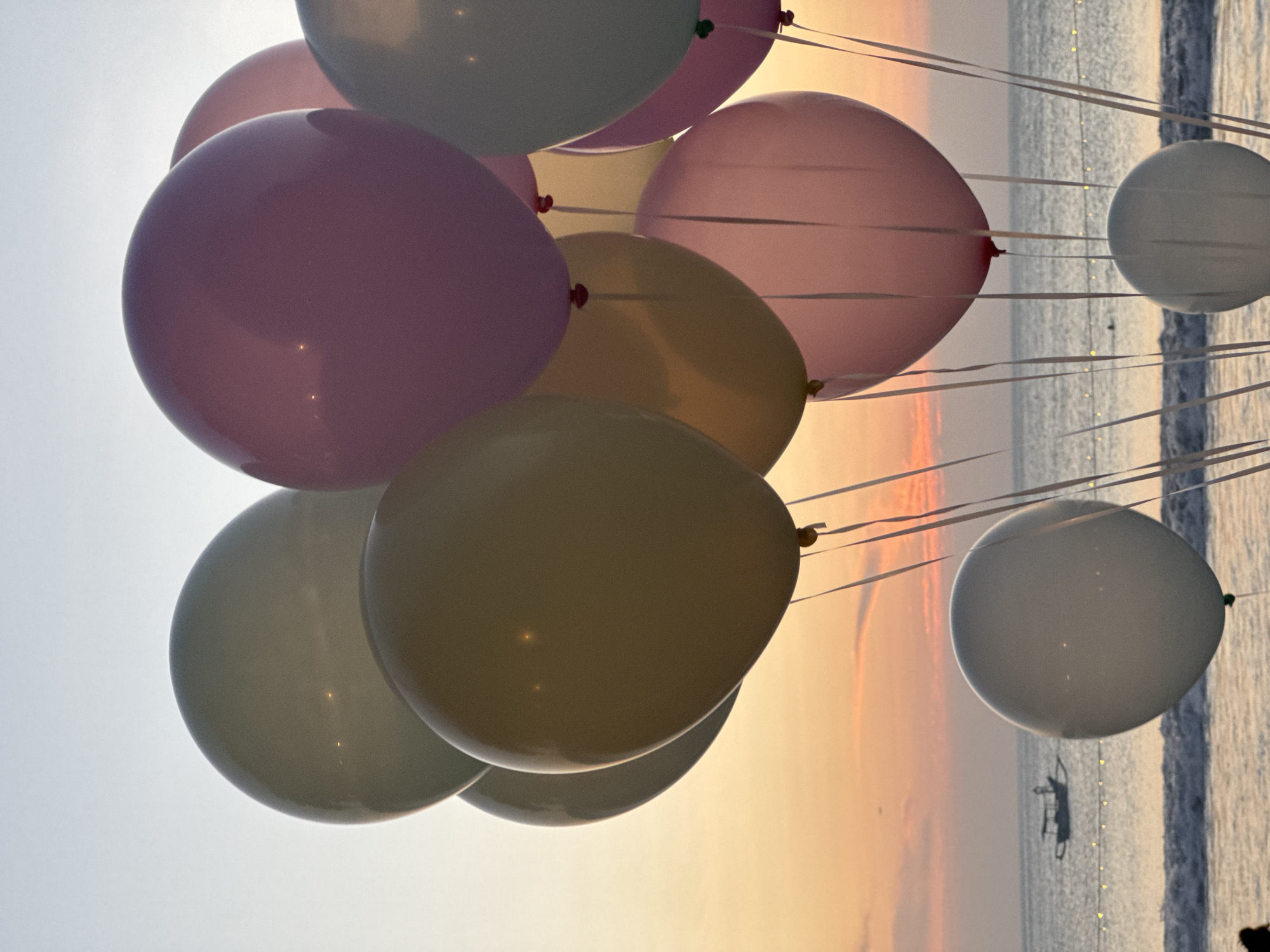 A cluster of pastel-colored balloons, including pale green, lavender, yellow, and pink, are tethered together, floating against a hazy sky at sunset. The balloons are the primary focus of the image. In the background, the ocean is visible with gentle waves breaking on a beach. A small fishing boat with a single figure on board is seen in the distance. A string of lights is visible along the shoreline. The overall scene suggests an outdoor celebration or event taking place at the beach in Kuta Selatan, Indonesia.