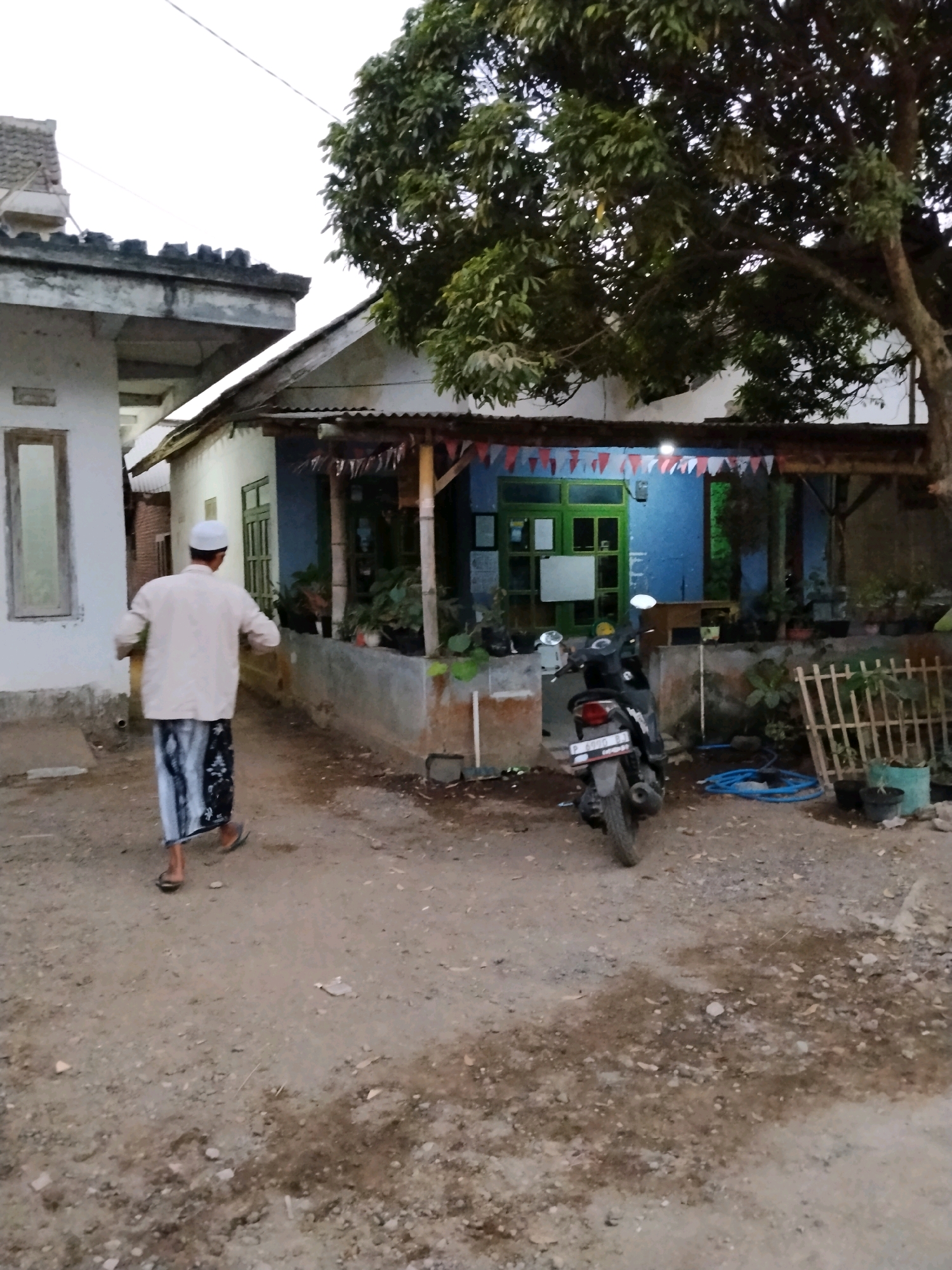 A man wearing a white skullcap and a light-colored long-sleeved shirt over a patterned sarong walks away from the camera on a dirt path in front of a residential building in Panggungan, Indonesia. He is wearing flip-flops. To his right, a motorcycle is parked in front of the house. The house has a blue-painted wall and a green-framed door with glass panes. A row of small triangular flags in red and white is strung across the front of the house under a corrugated metal roof extension. Plants in pots are visible on a ledge in front of the house, and a wooden fence stands to the right of the motorcycle. A large tree with dense foliage is to the right of the house, partially obscuring it from view. The ground is a mix of dirt, small stones, and fallen leaves. The sky is overcast.