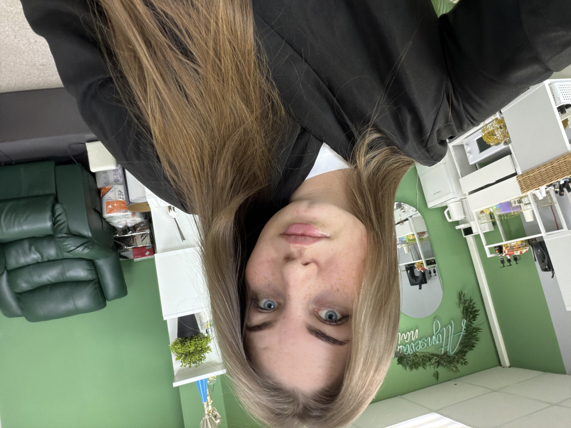 A woman with fair skin, blue eyes, and freckles is centered in the frame. She has long, brown hair that falls around her face and is wearing a black blazer over a white shirt. The background features a green wall with white shelves and cabinetry. On the left side of the frame is a green leather chair. A decorative circular sign with the words "Allyuriseva nai" in Russian is visible on the wall. This appears to be taken inside a business establishment, possibly a beauty salon in Chaikovsky, Russia.