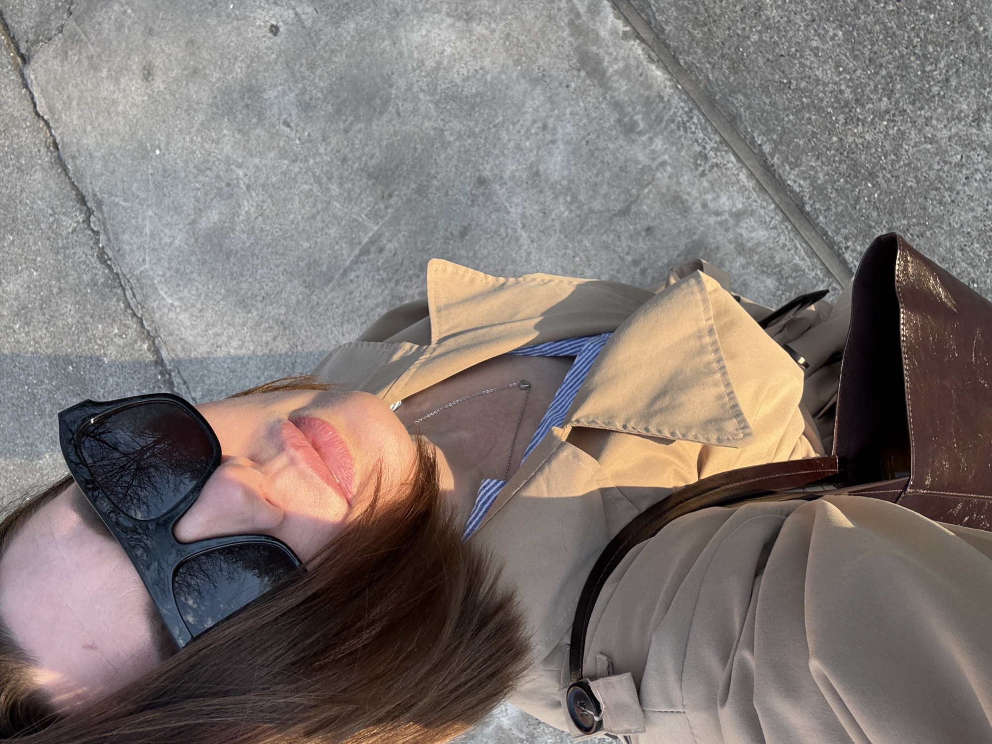 A photograph taken from an overhead perspective shows a woman in Chaikovsky, Russia. She is lying on a concrete surface outdoors, possibly a sidewalk. She is wearing sunglasses and a beige trench coat. A brown leather bag is positioned beside her. She is also wearing a silver necklace and a striped blue and white shirt underneath the trench coat. The shadows suggest the photo was taken during daylight hours. The surroundings are primarily urban, focusing on the concrete ground and the woman's attire and accessories.