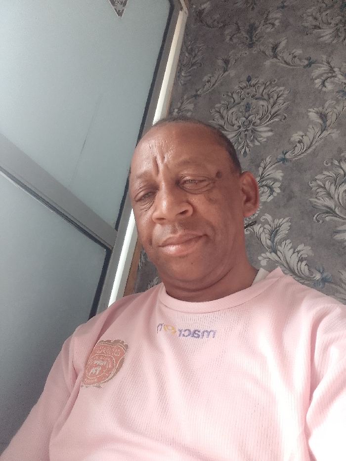 An adult man, likely of African descent and appearing middle-aged to elderly, is depicted in a close-up, head-and-shoulders shot. He has short, dark hair with a visible receding hairline and noticeable wrinkles on his forehead and around his eyes. He looks directly at the camera with a neutral to slightly content expression; his left eye (viewer's right) appears slightly less open than his right.
He is wearing a light pink, long-sleeved crew-neck shirt. On the left side of his chest (viewer's right), there is a circular or shield-shaped emblem in shades of orange, red, and gold, featuring an intricate design that may include text or a crest, though it is not clearly legible. Below the neckline on the right side of his chest (viewer's left), there is a mirrored text logo in purple or dark blue that appears to read "moyosom" or similar.
The background is divided into two distinct areas. To the left of the man, there is a smooth, light-colored surface, possibly part of a door or window frame, with a vertical dark line and a lighter, potentially reflective area further to the left. To the right, the wall is covered with a patterned wallpaper featuring a classic damask or baroque motif in various shades of grey and silver, accented with subtle light blue or teal within some of the decorative scrolls.
The lighting is soft and even, and the man appears to be taking a selfie or posing for a casual photograph indoors.