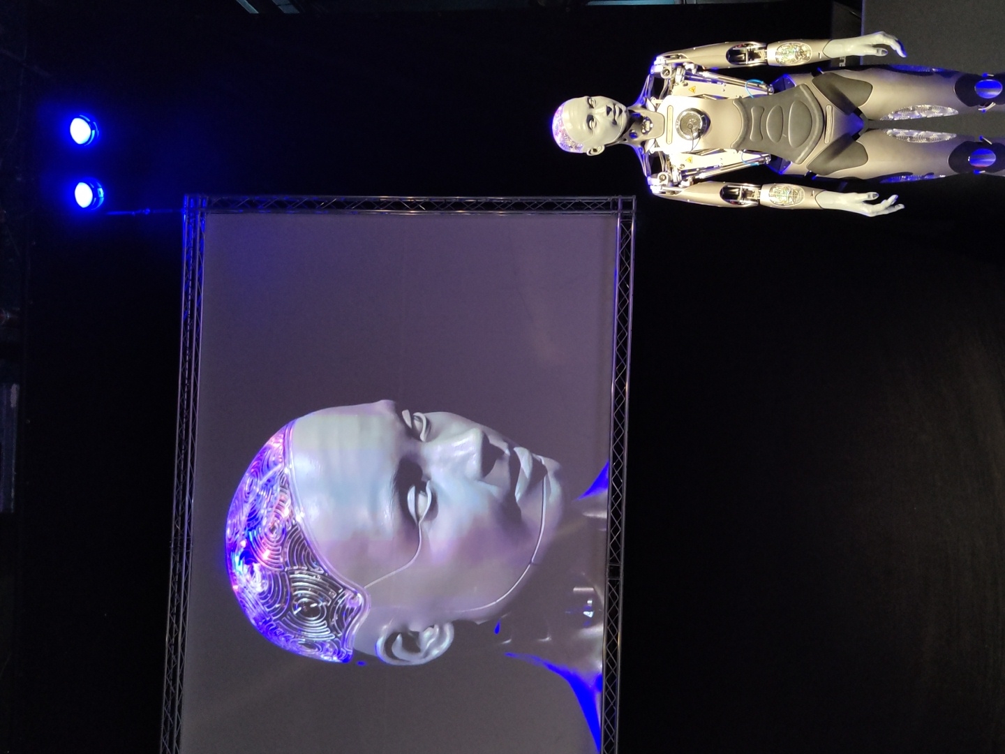 In a dimly lit indoor venue, likely a conference or exhibition hall, a large screen displays a close-up of a human-like robot's head. The robot's head on the screen is a pale gray, with intricate, glowing blue and purple circuits visible beneath a translucent skullcap. Its expression is neutral, and its gaze is directed slightly to the left.
In the foreground, to the right of the screen, stands a full-body, life-sized robot. This robot is also pale gray and silver, with visible mechanical joints and intricate detailing on its chest and abdomen. Its head is smooth and hairless, with a similarly neutral expression. The robot is standing upright, facing forward with its arms slightly away from its body.
The background is predominantly black, with the screen and the robot being the main sources of light. Two bright blue spotlights are visible above and to the right of the screen, casting a strong illumination. The overall impression is one of a futuristic presentation or demonstration. The venue appears to be spacious, with a dark ceiling and floor. The image does not provide any specific indicators of the city of Haifa, Israel.