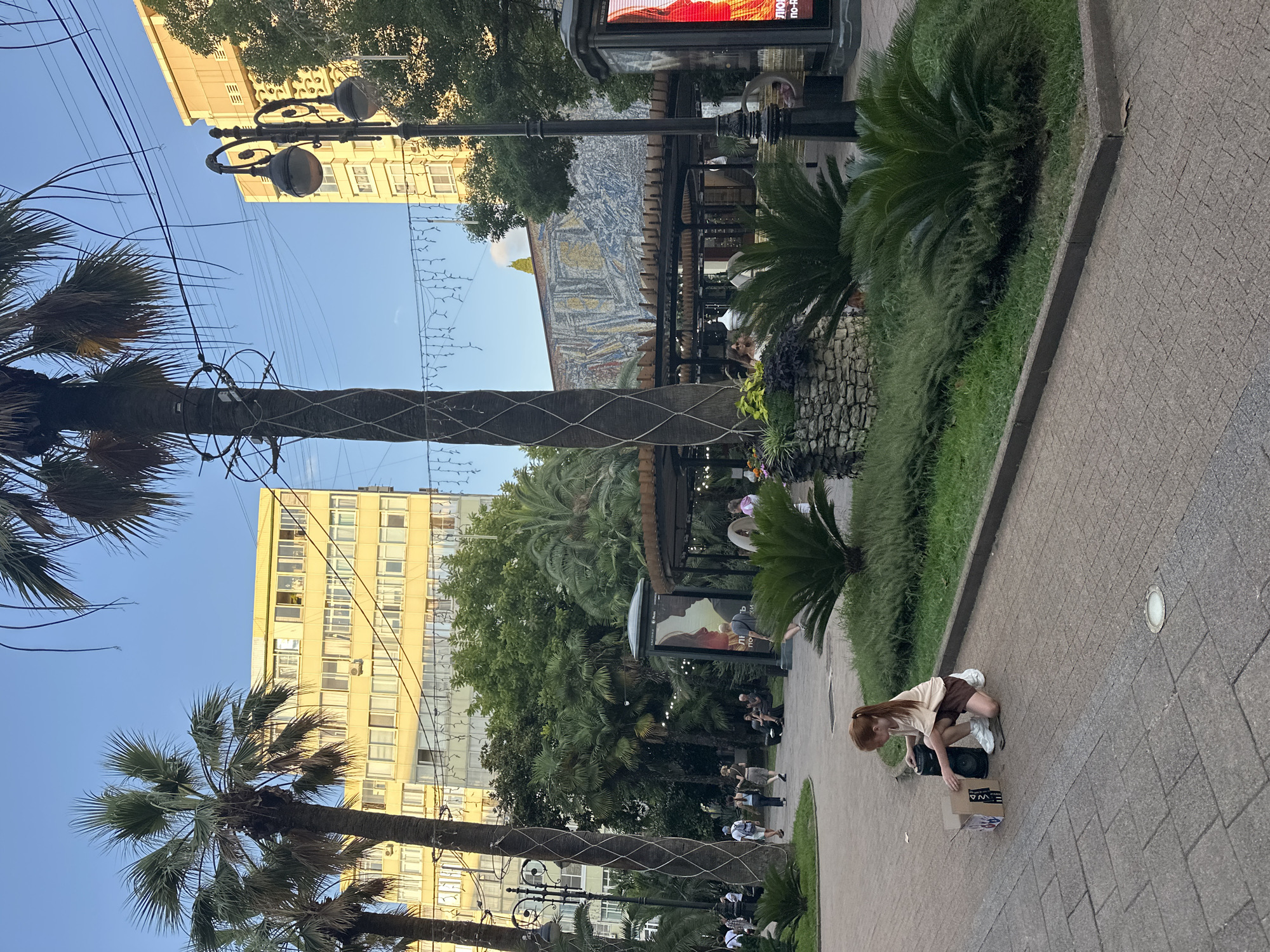 A young girl with red hair kneels on a brick walkway in an outdoor public space, interacting with a portable speaker and a cardboard box. She is wearing a tan t-shirt, brown shorts, and white sneakers. Palm trees and lush green plants surround the area, with a large, yellow building visible in the background. The sky is clear and blue. In the mid-ground, other people are seen walking and sitting, and a mosaic mural adorns the side of a building. The scene appears to be a park or pedestrian area in Sochi, Russia, given the presence of palm trees and the architectural style.