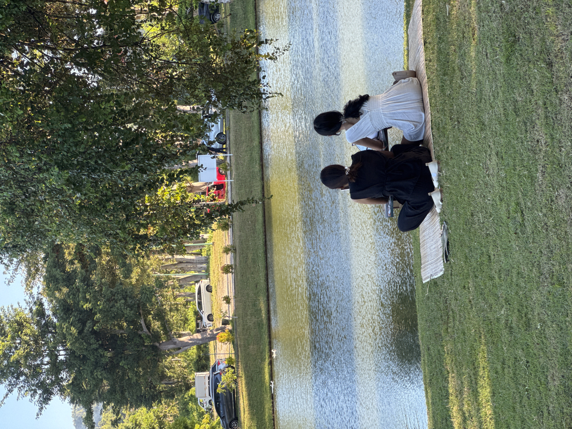 Two people sit on a woven mat on the green grass next to a body of water. One person is wearing a white dress and the other a black dress. A tree with thick foliage is to the left of the frame. Across the body of water, cars and a red vehicle are visible, suggesting an adjacent road. Based on the environment, the location is likely in Kuta Selatan, Indonesia, given the request.
