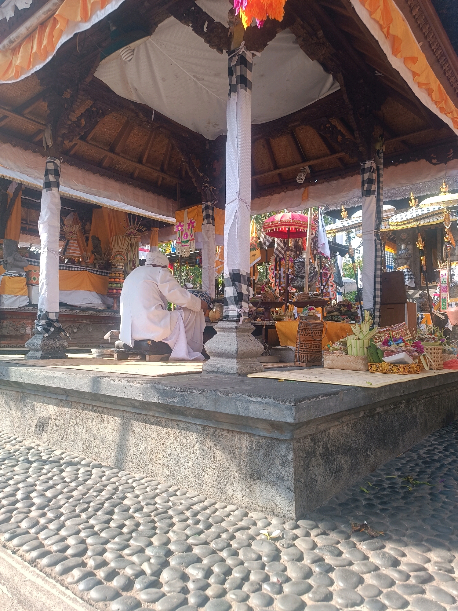 The image shows a man in a white robe and hat kneeling on a dark mat in front of an altar. The altar is decorated with yellow and orange fabric, offerings of food, and what appear to be religious statues. The scene is set outdoors, under a thatched-roof pavilion supported by stone columns wrapped in black and white checkered fabric. The ground is paved with smooth, grey stones. In the background, more of the temple complex is visible, including ornate carvings and flags. The overall impression is that of a religious ceremony taking place in a Balinese temple, likely in the city of Tabanan, Indonesia, given the architectural style and cultural elements.