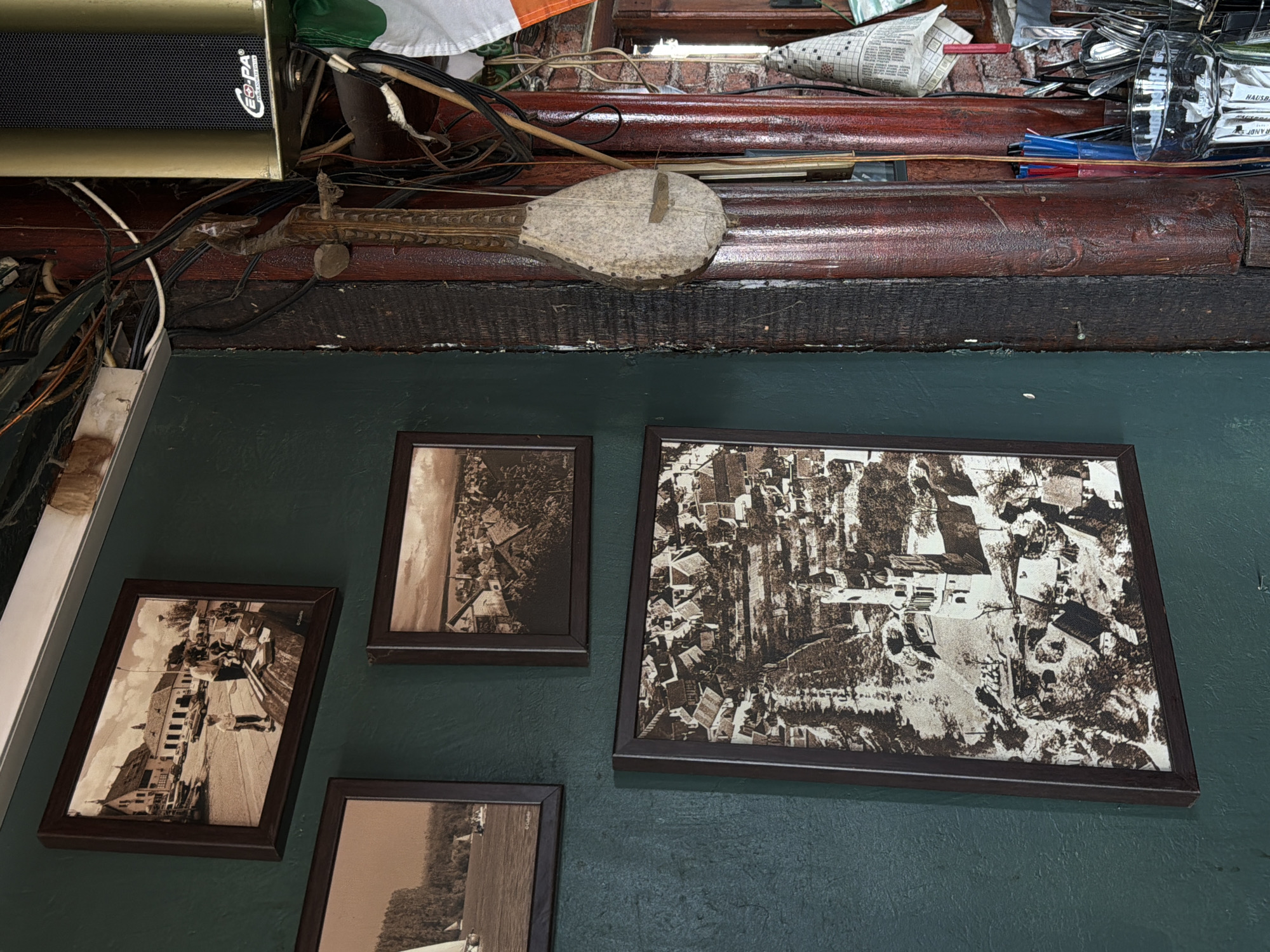 The image depicts a wall with a collection of framed photographs. The wall is painted green and the frames are dark brown. The photos appear to be in sepia tones and depict urban scenes, possibly of a historical nature. Above the photographs, there is a cluttered shelf with various objects including a speaker, what looks like a bellows, newspapers, utensils, and other miscellaneous items. The scene does not clearly identify a specific location in Palilula Urban Municipality, Serbia, and further information would be needed to determine a location.