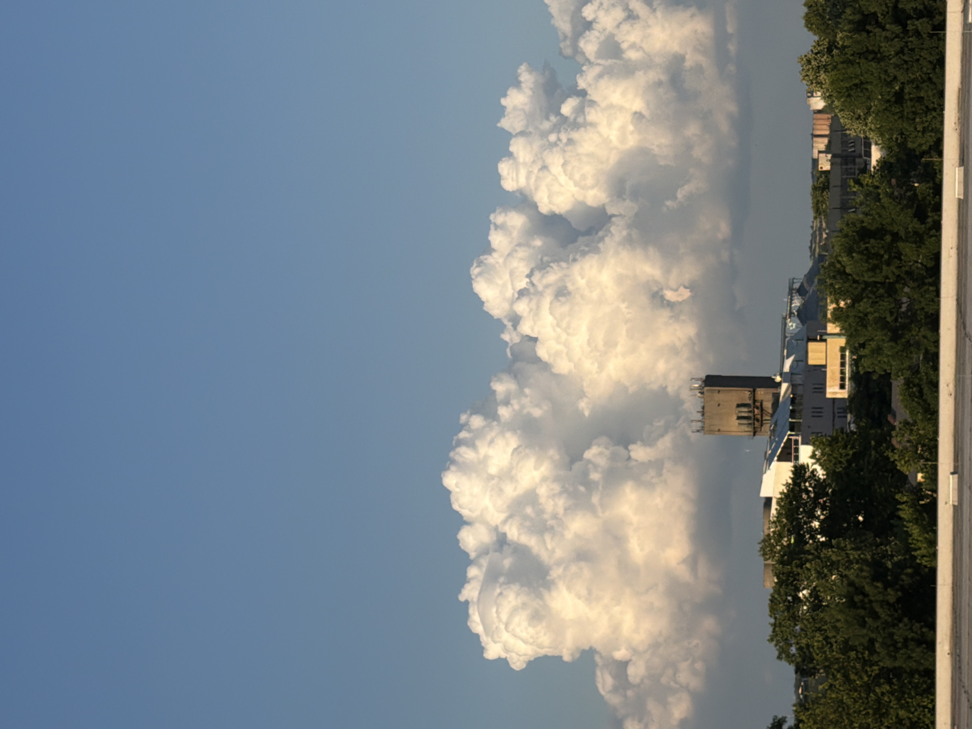 The image presents a panoramic view of an urban-industrial landscape set against a dynamic sky. Approximately the top 40% of the frame is dominated by a clear, uninterrupted blue sky, transitioning from a slightly darker shade at the apex to a lighter hue towards the horizon.
Below this, a vast formation of dense, white cumulus clouds stretches horizontally across the entire width of the mid-ground. These clouds exhibit significant vertical development and are intensely illuminated by warm, possibly late-afternoon sunlight from the right, creating bright white highlights and distinct golden-orange shadows on their undulating surfaces.
Beneath the cloud base, a collection of buildings and structures forms a distant skyline, partially obscured by a dense canopy of dark green deciduous trees that occupies the lower mid-ground. Prominently centered among these structures is a tall, dark grey, rectangular concrete tower, possibly industrial or utilitarian, with visible antennae or equipment on its flat roof. Surrounding it are various lower-rise buildings, predominantly in shades of white, light grey, and beige, with some showing flat or subtly sloped roofs. Hints of industrial architecture, including what may be storage silos, are visible on the far left.
The immediate foreground consists of a flat, light grey paved or concrete surface, likely a rooftop or elevated platform, featuring subtle linear markings and a few small, rectangular white objects. The overall lighting suggests late afternoon, characterized by strong, directional light and well-defined shadows. No human figures or discernible activities are present in the scene.