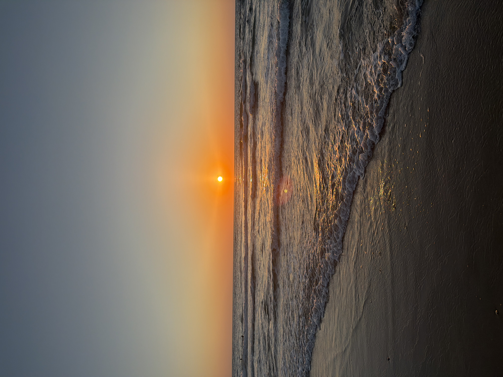The image depicts a sunset over the Mediterranean Sea at Bat Yam, Israel. The sun, a bright orange disc, is positioned near the horizon, casting a warm glow across the sky and water. The sky transitions from a pale blue at the top to a vibrant orange near the horizon. Gentle waves are rolling towards the shore, creating white foam as they break. The wet sand in the foreground reflects the sunset colors. A few indistinct figures are visible in the water, suggesting people might be swimming or wading. The overall scene is tranquil and serene, characteristic of a beach at dusk.