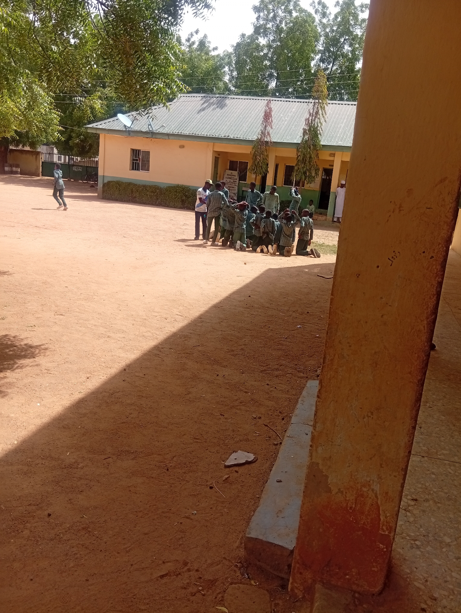 In Dutse, Nigeria, a group of young students, dressed in green and white uniforms, are kneeling on a dusty, reddish-brown courtyard. The students are gathered in front of a light yellow, single-story building with a green metal roof. Several trees with lush green foliage surround the building and the courtyard, casting shadows across the ground. A tall, roughly textured, tan-colored pillar occupies the foreground on the right side of the image, obscuring part of the building and the scene. The pillar has some markings on its surface. In the background, the building appears to have multiple windows and a covered walkway. Other individuals, possibly teachers or older students, are standing near the building. One person in a white garment stands to the right of the building. The overall setting suggests an outdoor area associated with a school or educational institution.