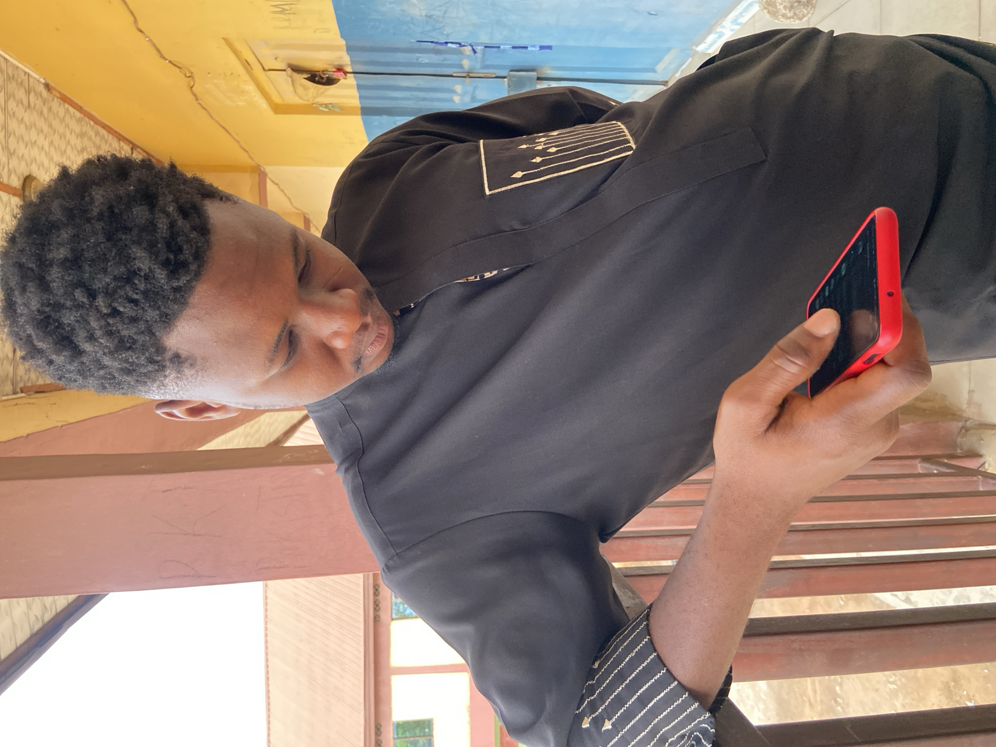 A dark-skinned man with short, curly black hair is holding a red smartphone. He is wearing a black outfit with white stripes on the sleeves and is standing outdoors. The location appears to be Tudun Wada Bibinu, Nigeria based on the environment. The background contains elements of a building facade and outdoor space. The man seems to be looking at or interacting with the phone. The surface he stands on has wooden bars.