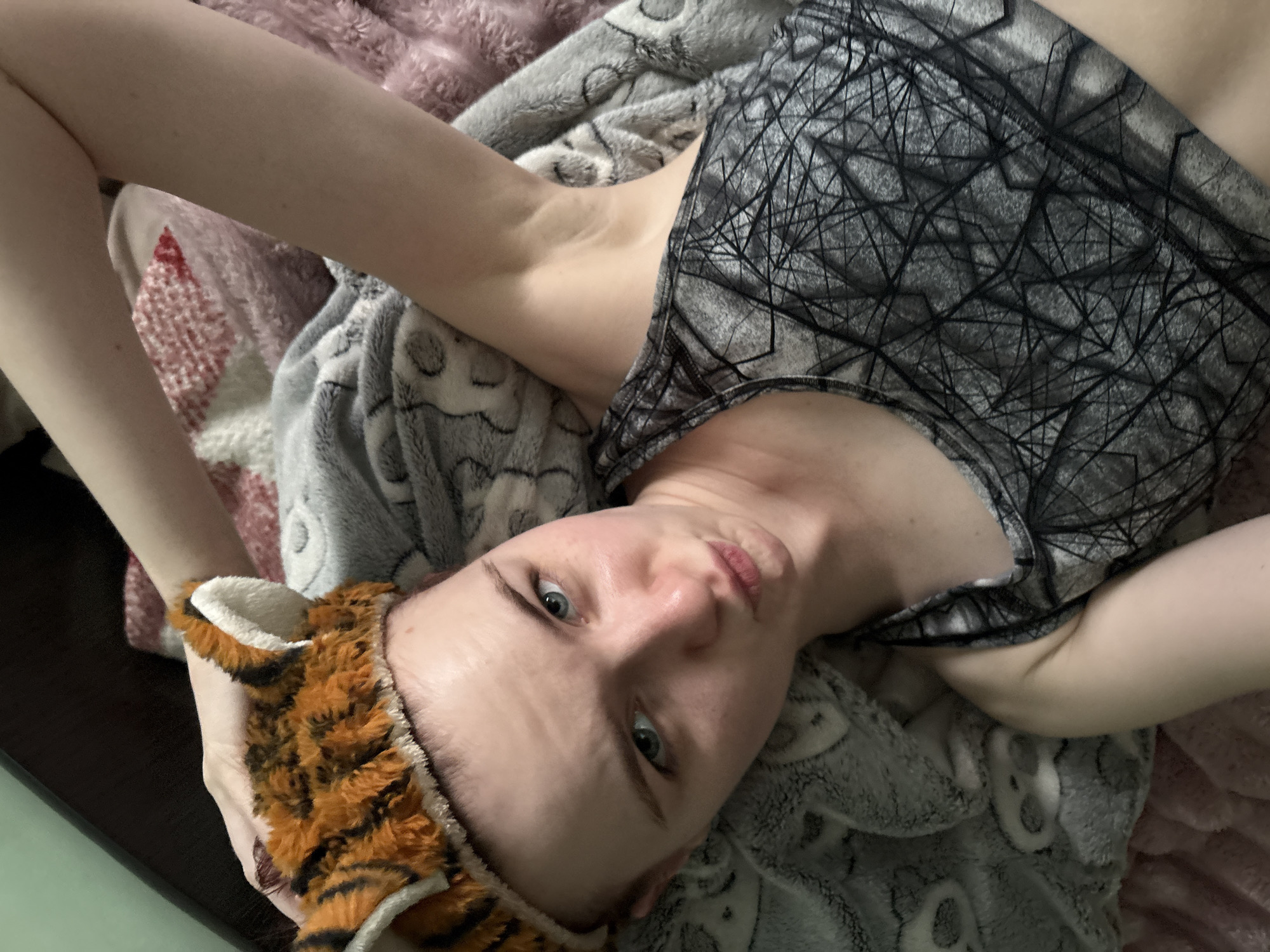 A person with light skin and blue eyes is lying on their back on a bed covered with blankets. The person is wearing a grey tank top with a geometric pattern and a tiger-eared headband. Their arms are raised above their head. The blankets are grey and pink, and one has a pattern of cartoon animals. The lighting is dim, suggesting it could be nighttime or a dimly lit room. The person's expression is neutral, with their gaze directed towards the camera. There is no visible text in the image.