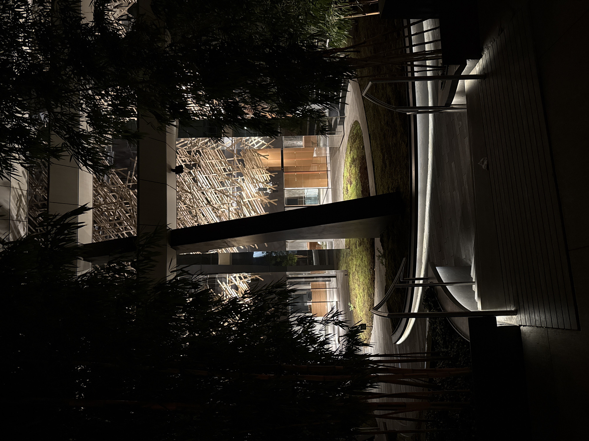 At night, a low-angle shot captures the architecture of a building in Vancouver, Canada, partially obscured by foliage. The modern building features stacked levels with glass windows, revealing internal wooden structures or artwork. An illuminated walkway runs along the building's exterior, marked by sleek metal railings and contrasting dark shadows. Vegetation flanks the walkway, and the shot is taken from a dark, obscured position, adding depth and mystery to the scene.