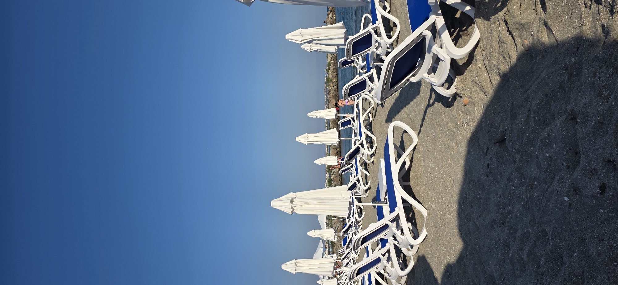 The image shows a beach in Pomorie, Bulgaria. The beach is covered in dark sand. There are many white beach chairs with blue cushions and closed white umbrellas lined up on the sand. In the background, there is the sea and a rocky breakwater. A few people are visible in the water and on the breakwater. The sky is clear and blue. The scene appears to be set in the late afternoon or early evening, as indicated by the long shadows cast by the beach chairs.