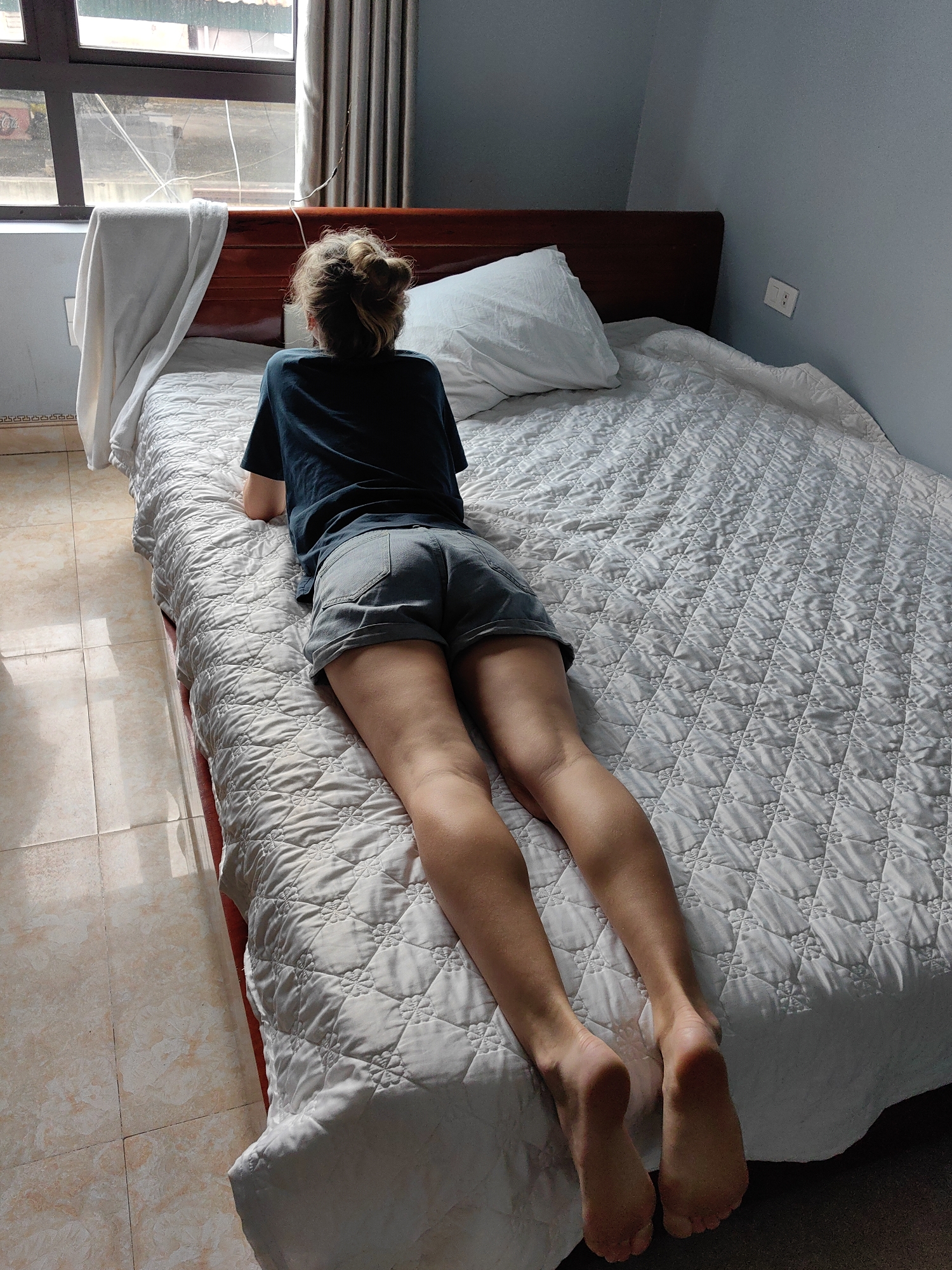 A person is lying prone on a bed in a room. The individual is wearing a dark blue t-shirt and grey shorts. Their hair is gathered in a bun. A white pillow rests on the head of the bed. The bed is covered with a white quilted bedspread. A window with white curtains is on the left. The floor is tiled. A white towel hangs off the side of the bed. The image was taken in Bai Chay Ward, Vietnam.