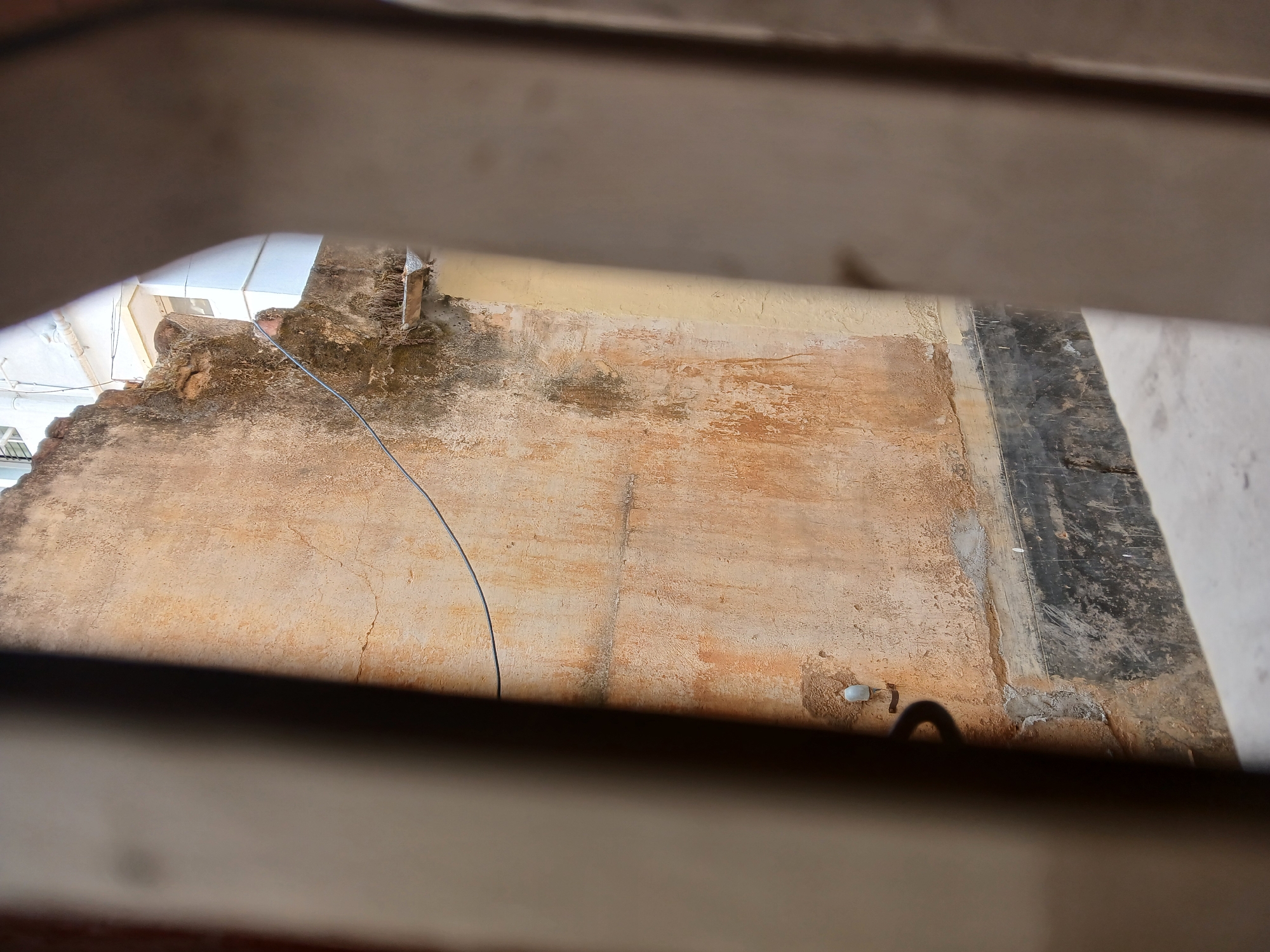 The image shows a view of an exterior wall in Bengaluru, India, as seen through a partially obstructed opening, possibly a window or vent. The wall is predominantly a weathered, textured surface with a patchwork of tan and beige tones, suggesting repairs or uneven aging. There's evidence of water damage or discoloration in areas, particularly near the top. A wire runs diagonally across the wall's surface. To the right, there is a dark, possibly painted or sealed, section of the wall. In the lower right corner, there is a drainpipe with a small cap or plug. The image is framed by blurred structural elements of the viewing aperture.