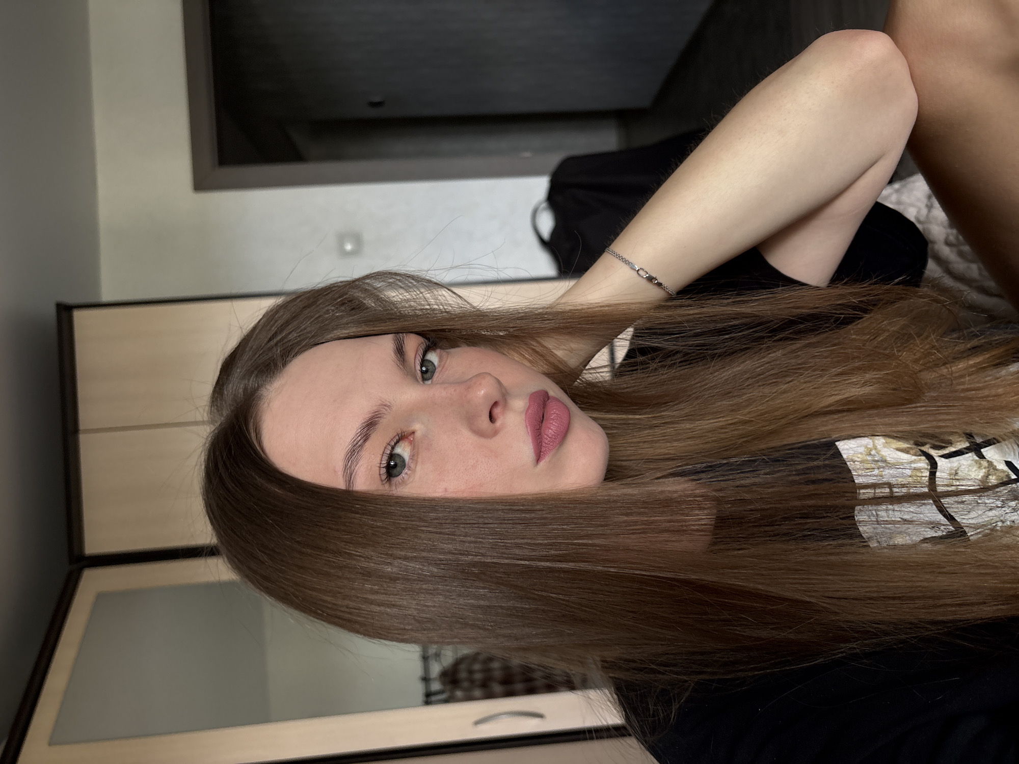A young woman with long, brown hair and light blue eyes is pictured in a selfie. She is wearing a black t-shirt and has light pink lipstick on. She is resting her head on her hand, which is adorned with a silver bracelet. In the background, there is a closet with mirrored doors and a doorway leading to another room. The setting appears to be a bedroom within an apartment. The image was taken in Perm, Russia.