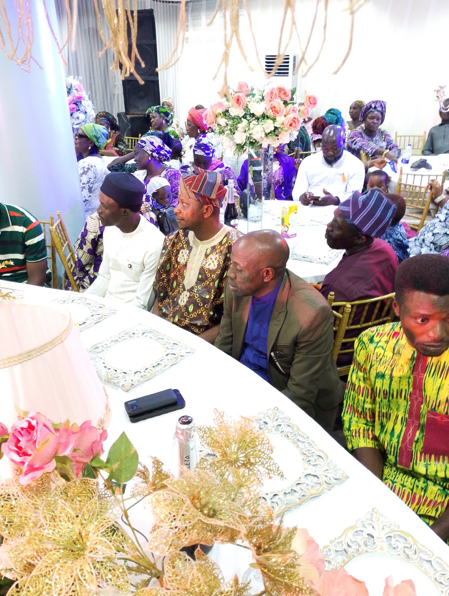 The image shows a gathering of people in a large room, likely a banquet hall, possibly located in Kubwa, Nigeria. The room is decorated for an event, with tables covered in white cloths and adorned with floral arrangements (roses and other flowers). Several people are seated at the tables, dressed in traditional attire, including head wraps (gele) and colorful patterned shirts. A man is wearing a brown suit jacket, and another wears a traditional Yoruba fila cap. There are floral decorations, frames, and a phone on one of the tables. Behind the tables, more people are seated, and decorations hang from the ceiling. A speaker and air conditioner unit are visible in the background. The mood seems to be one of formal celebration or gathering.