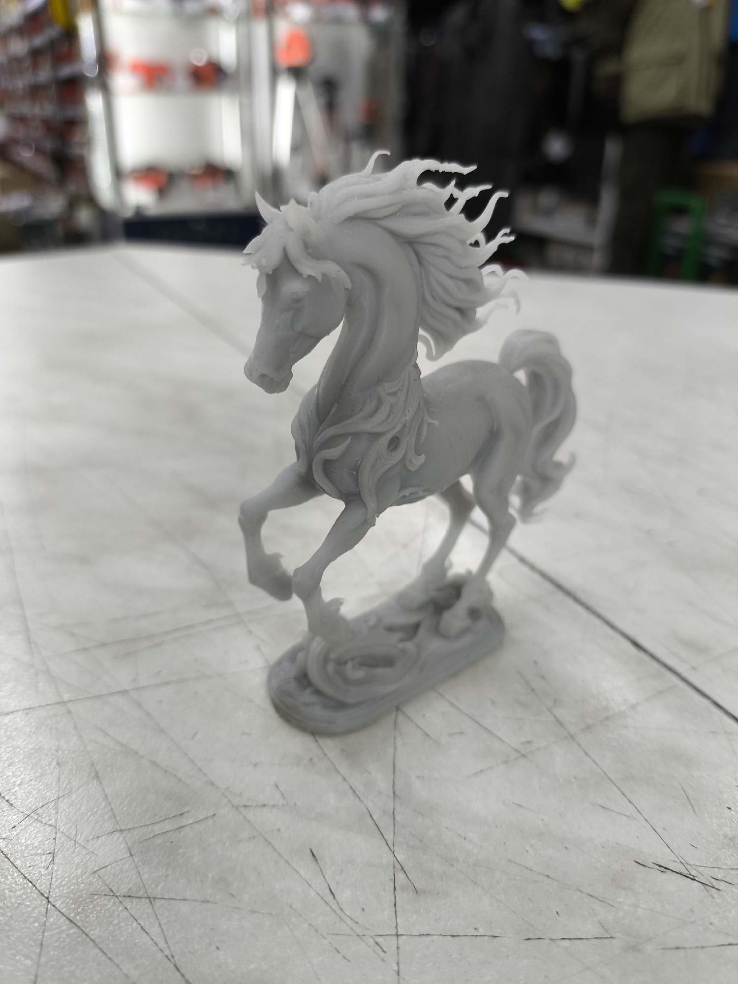 A light gray, three-dimensional model of a galloping horse is positioned on a white, textured surface that appears to be a table or workbench. The horse is depicted in mid-stride with its head turned and mane flowing. The surface has visible lines and some scuff marks. In the blurred background, there are shelves with various items, some appearing to be red and white. The overall environment suggests an indoor setting, possibly a workshop or store. The location is identified as Solikamsk, Russia. There are no people clearly visible in the immediate foreground or interacting with the horse model.