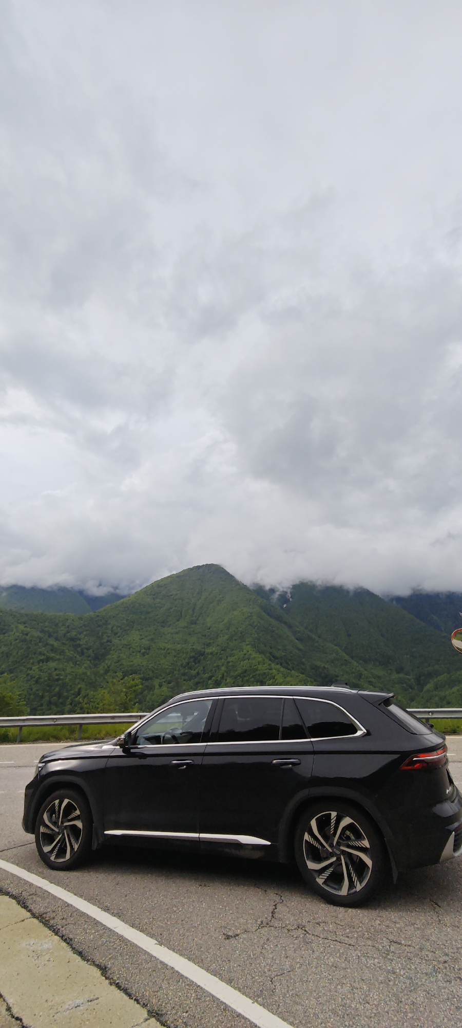A black SUV is parked on the shoulder of a road. Behind the car is a landscape of forested mountains partially obscured by clouds. The sky is overcast. The location is Chvizhepse, Russia.