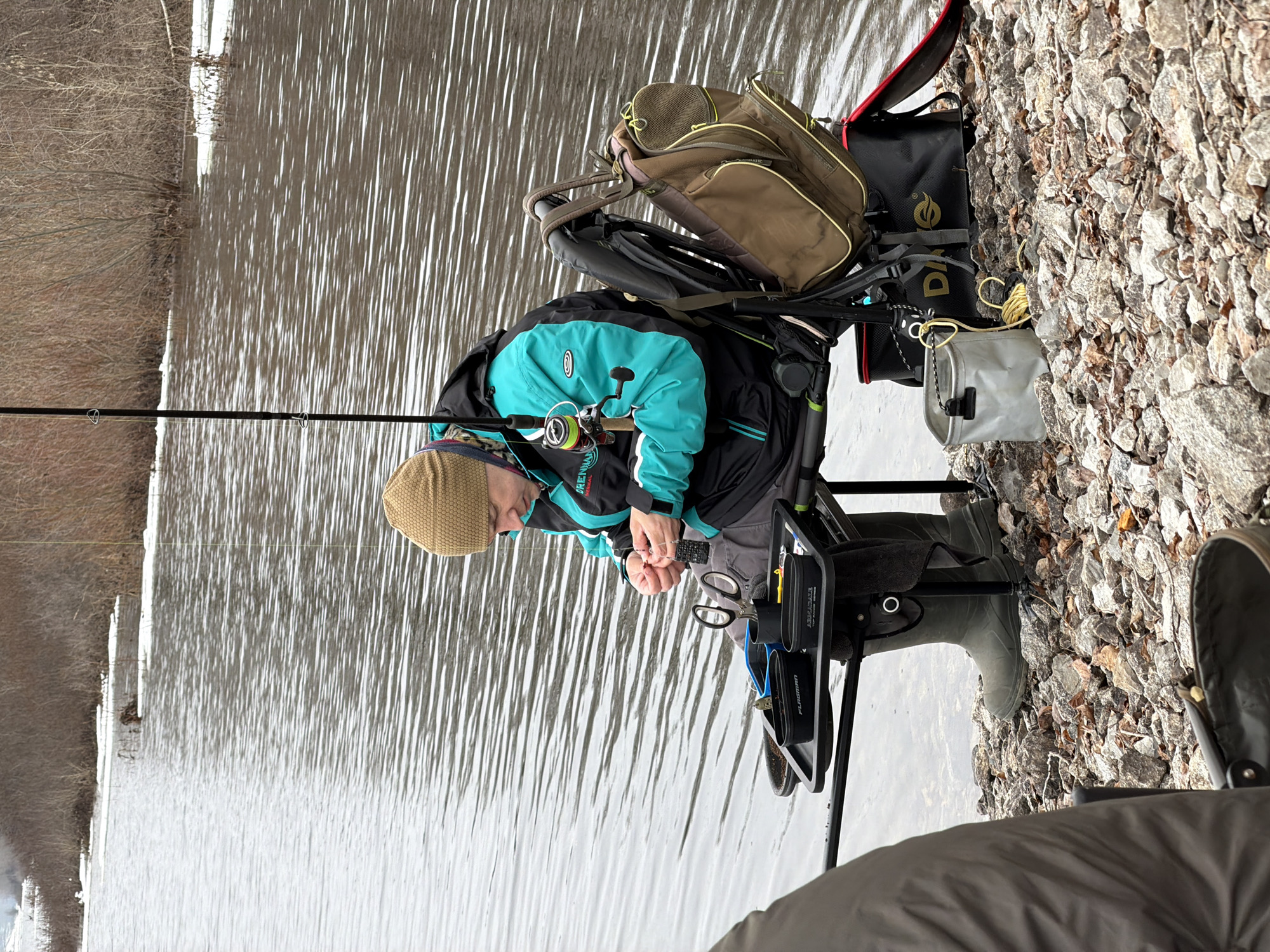 A man is fishing on a cloudy day, possibly in the city of Chaikovsky, Russia. The man, wearing a beige knit hat and a blue and black jacket, is seated on a specialized fishing chair that is equipped with accessories. He is positioned next to the water, with a fishing rod extending into the water. The chair is loaded with a brown backpack, a black bag, and a gray pouch. He appears to be adjusting something in his hands. The ground consists of a rocky, pebble-covered shoreline. The water has ripples and reflects the overcast sky. Bare trees line the opposite shore. The photo is taken at an angle, with the man and his equipment positioned to the right side of the frame.