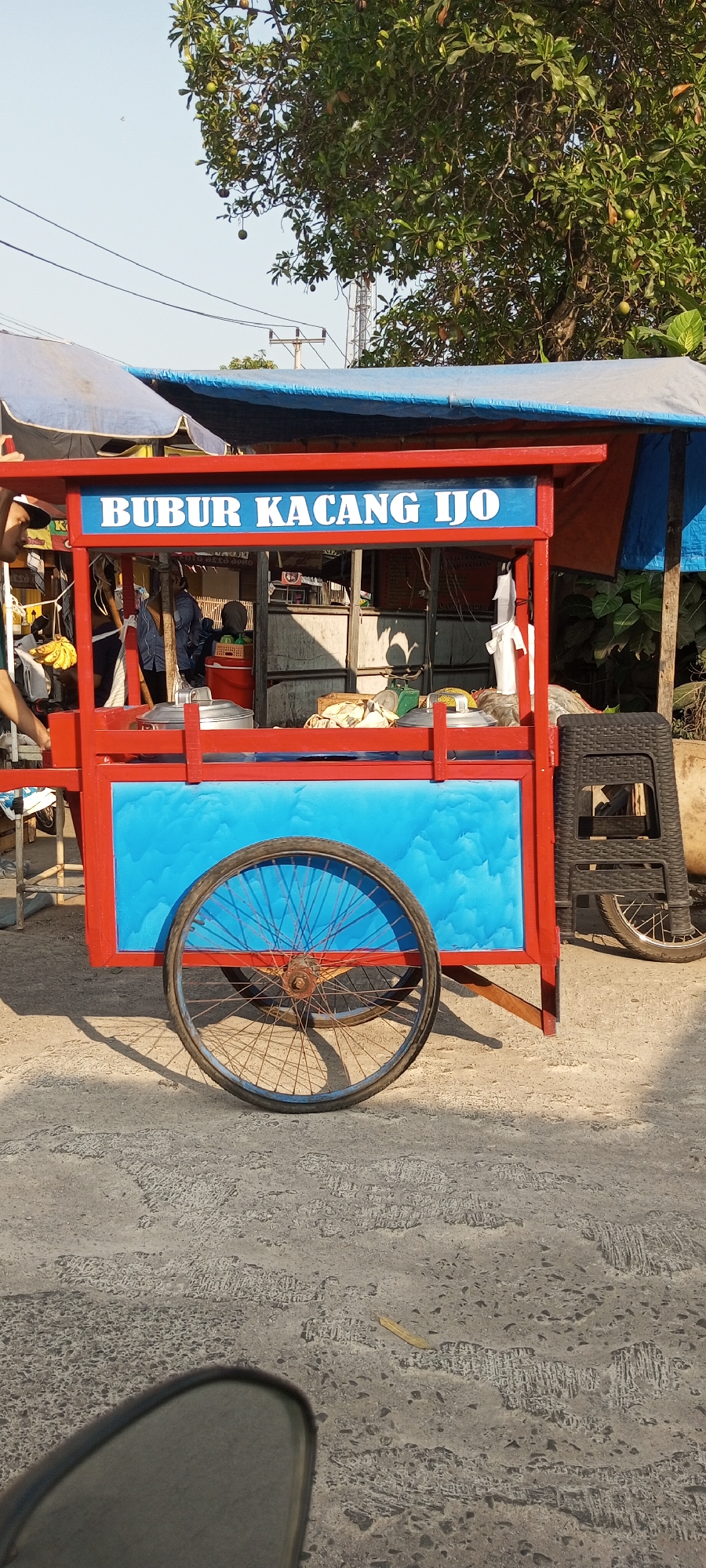 The image shows a food cart in Setu, Indonesia. The cart is red and blue, with "BUBUR KACANG IJO" written in white letters on a blue background at the top. There are two wheels visible, one larger and one smaller, on the cart. In the background, there are trees, power lines, and other stalls. A person is partially visible on the left side of the cart, and a black plastic chair is on the right. The ground is paved with concrete.