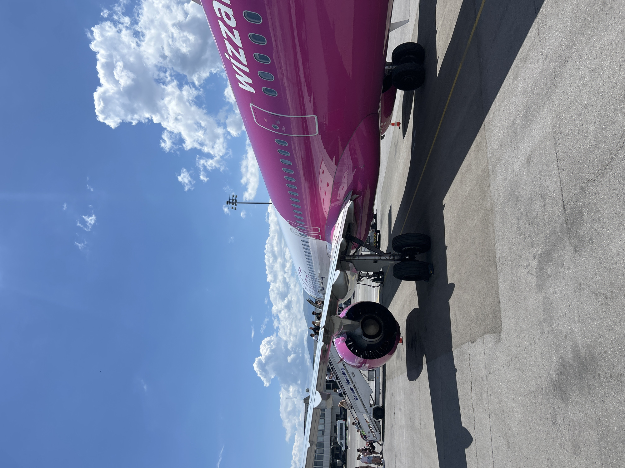 A bright pink Wizz Air Airbus A320 aircraft is parked on the tarmac at Sofia Airport in Sofia, Bulgaria, under a partly cloudy blue sky. Passengers are boarding the plane via a mobile stairway positioned next to the left wing, near the engine. The aircraft's livery features the Wizz Air logo in white text on the rear fuselage. In the background, a terminal building, a white van, and distant mountains are visible. The ground is asphalt, with a yellow line marking a boundary.