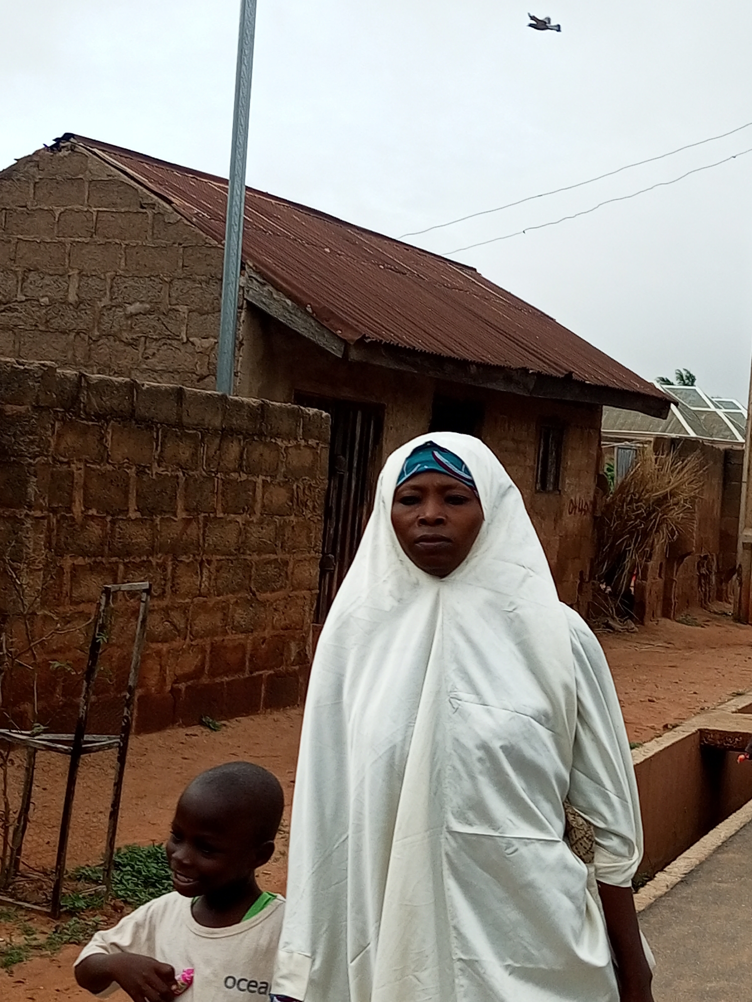 A woman wearing a white hijab and a blue headscarf stands in the foreground, looking forward. To her left, a young child in a white t-shirt looks to the left and smiles. Behind them, a rustic, brick building with a corrugated metal roof is visible. The ground is dirt, and there are some plants and a metal structure to the left of the child. In the sky, a bird is in flight. The overall environment appears to be a rural or semi-rural area.