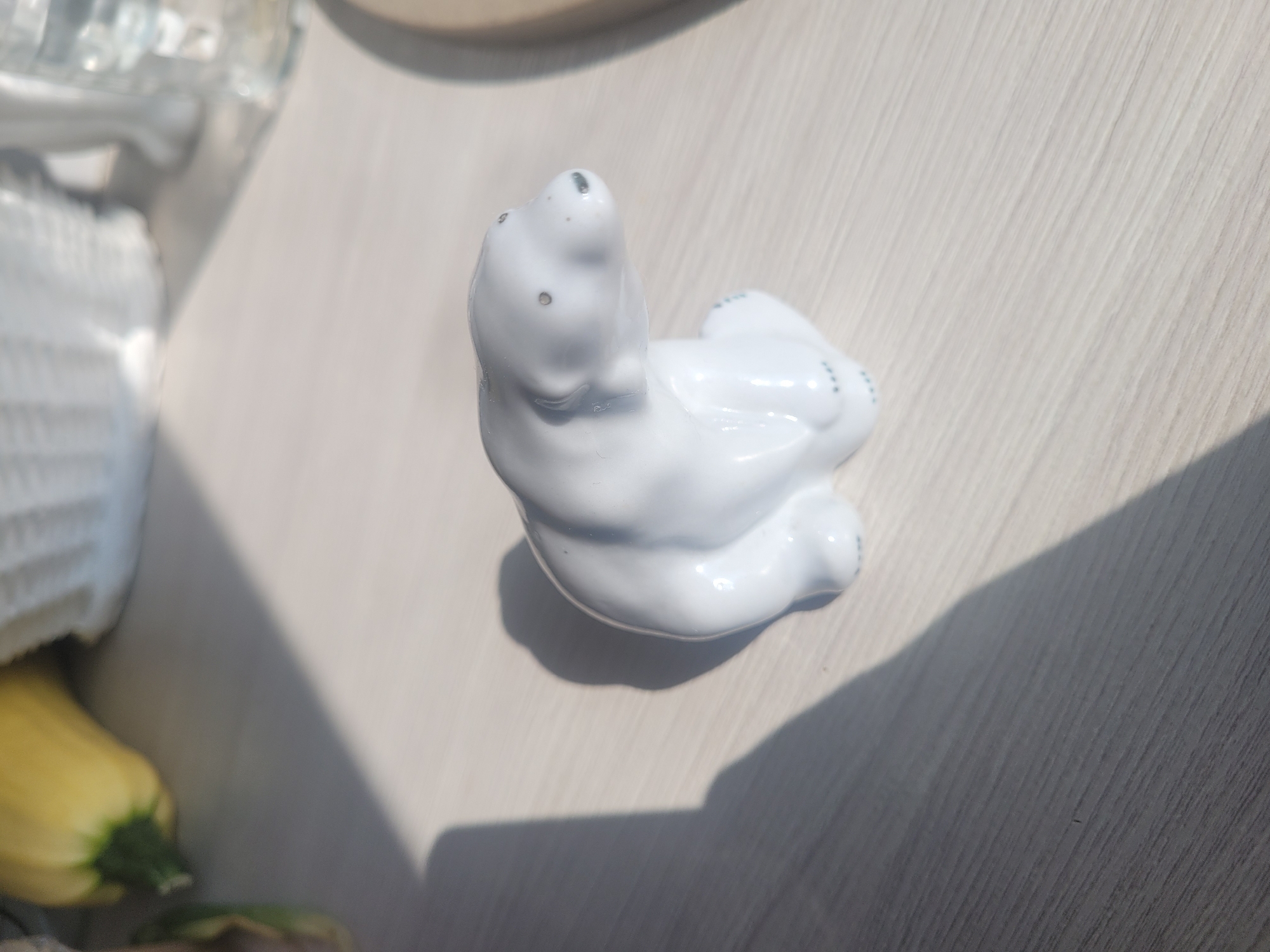 A white ceramic polar bear figurine sits on a light-colored, wooden surface. The surface has a wood grain texture, and is illuminated by sunlight, casting a prominent shadow of the figurine and another object to the left of the frame. In the background, out of focus, are what appear to be white woven baskets and a yellow vegetable, possibly a zucchini or squash. A clear glass is also partially visible in the background. The scene is lit by natural light, creating bright highlights on the figurine. The location is not specified, but it is a still life arrangement indoors, likely on a table or shelf.