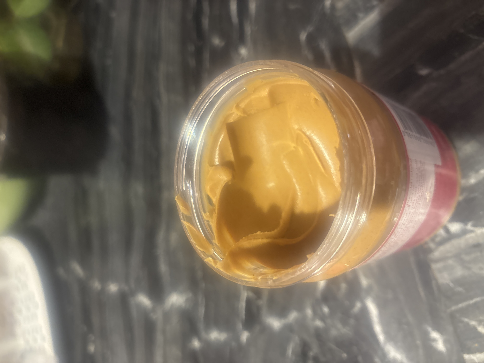 The image shows an open jar of peanut butter sitting on a dark, speckled countertop. The peanut butter is a smooth, creamy texture and fills the jar. The jar is clear plastic with a pink and white label around its base. The background is out of focus, but appears to be a kitchen environment with a hint of green foliage and dark shelving. There are no people or animals visible in the image. The location is not specified.