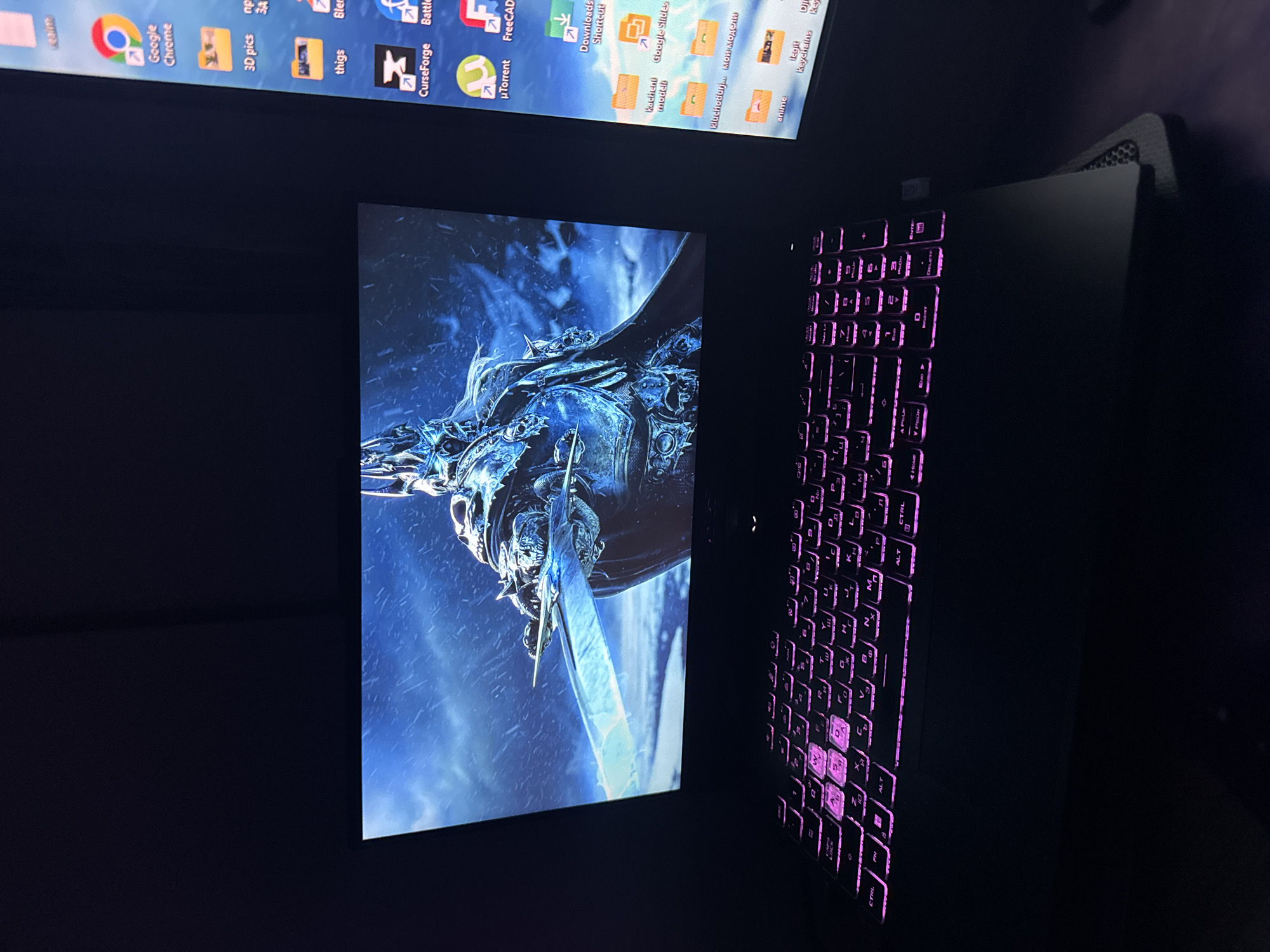 A black laptop is positioned on a dark surface, with its screen displaying an image of a character with a blue-toned armor and sword against a cloudy sky. The keyboard of the laptop is backlit with a magenta-purple hue, highlighting the keys. Above the laptop screen, a monitor shows a desktop with various icons of applications and folders, including Google Chrome, 3D pics, things, CurseForge, Battle.net, µTorrent, FreeCAD, Downloads Shortcut, Google Slides, and folders labeled "Kacheni modeli," "kluchodurj...Mой модели," and "anime," along with icons for "Dj Keychains."