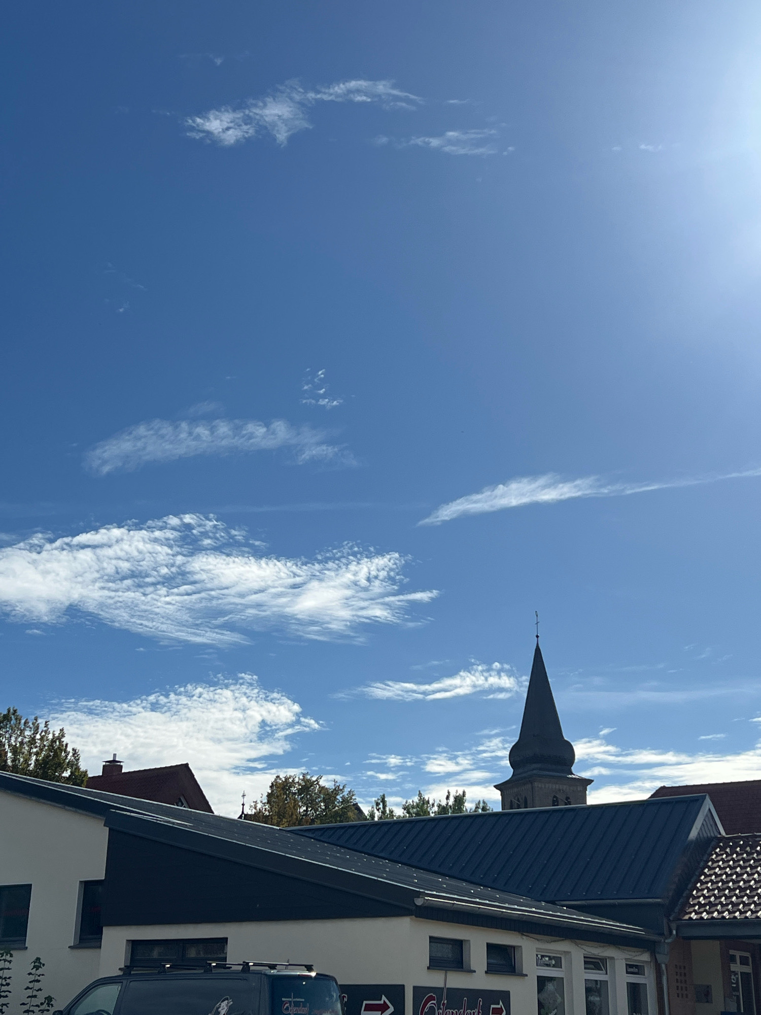 The image depicts a bright, clear sky with scattered white clouds, above a collection of buildings. A prominent feature is a dark, pointed church steeple, topped with a cross, rising from behind the rooftops. The buildings appear to be a mix of residential and commercial structures, with dark metal roofs and white facades. Some greenery and trees are visible between the buildings, suggesting a suburban or town setting. The lighting indicates it is daytime, with the sun positioned in the upper right portion of the frame, creating a slight glare. A dark-colored van with roof racks is parked in front of one of the buildings. There are no people visible in the frame.