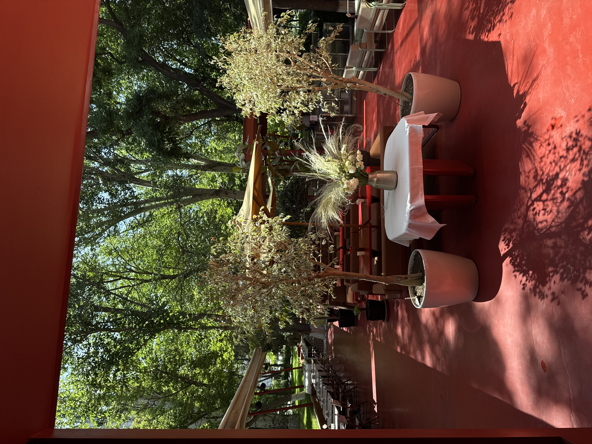 This image shows an outdoor seating area of a restaurant or cafe, likely in Yerevan, Armenia, given the architectural style and lush greenery common in the city's parks and public spaces. The foreground features a vibrant red-colored floor, bathed in dappled sunlight that casts intricate tree shadow patterns. Two potted trees, one on the left and one on the right, flank a table set with a white tablecloth. On the table, a metallic vase holds a bouquet of white flowers and decorative grasses. In the background, rows of tables and seating are arranged under awnings, with more trees and foliage creating a natural canopy. The overall atmosphere is serene and inviting, suggesting a pleasant dining experience amidst nature.