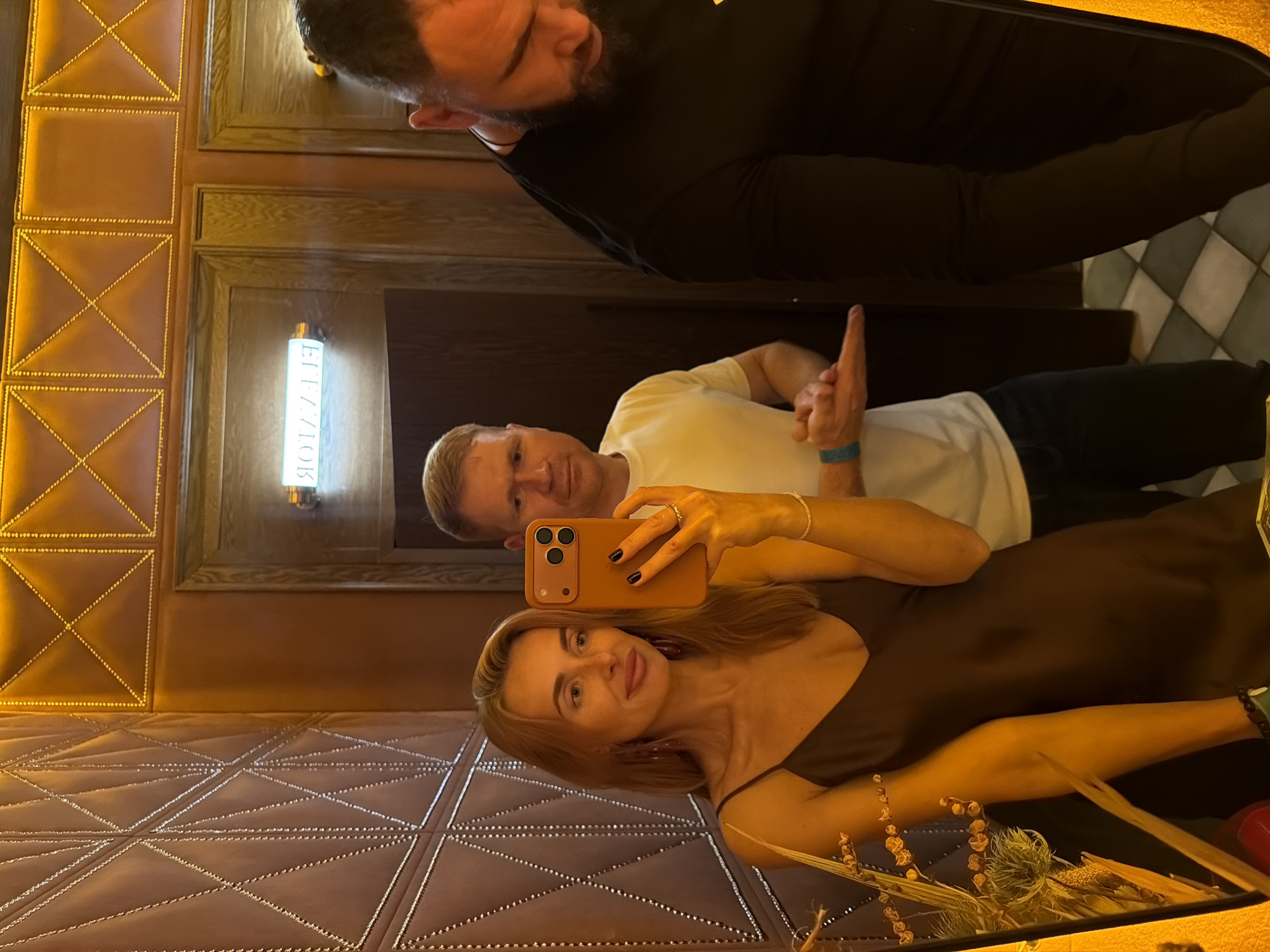 The image depicts three individuals taking a selfie in what appears to be a restaurant or bar in Krasnoyarsk, Russia. The woman in the foreground is holding a smartphone and facing the camera. She has blonde hair, light skin, and is wearing a brown dress. Behind her and to the left is a man with light skin and short blonde hair, wearing a white T-shirt. Above them, in the upper center of the image, a third man with dark hair and a beard is partially visible wearing a black long-sleeved shirt.
The environment includes decorative elements such as patterned upholstery and an illuminated sign that possibly says "ELEVATOR". The lighting is warm, creating a subdued ambiance. There are also floral decorations visible in the foreground. The floor tiles are visible in the background.