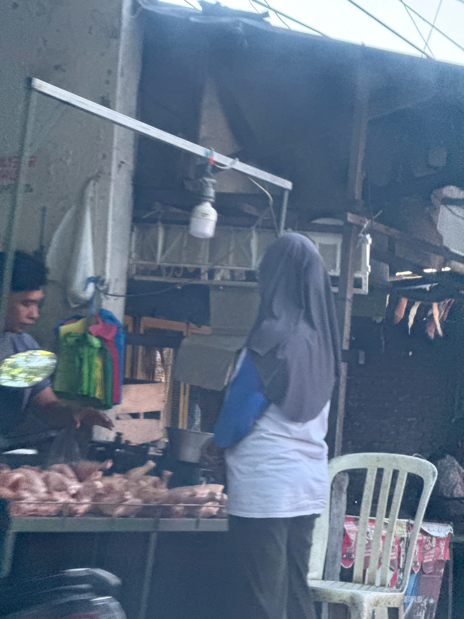 The image depicts an outdoor market stall in Surabaya, Indonesia. A vendor, partially visible on the left, is attending to a display of raw chickens on a metal rack. To the right, another person wearing a headscarf and a light-colored top stands. A white plastic chair is placed near the stall. The background shows the typical infrastructure of an open-air market, with various structures and possibly other vendors. The lighting suggests it is daytime.
