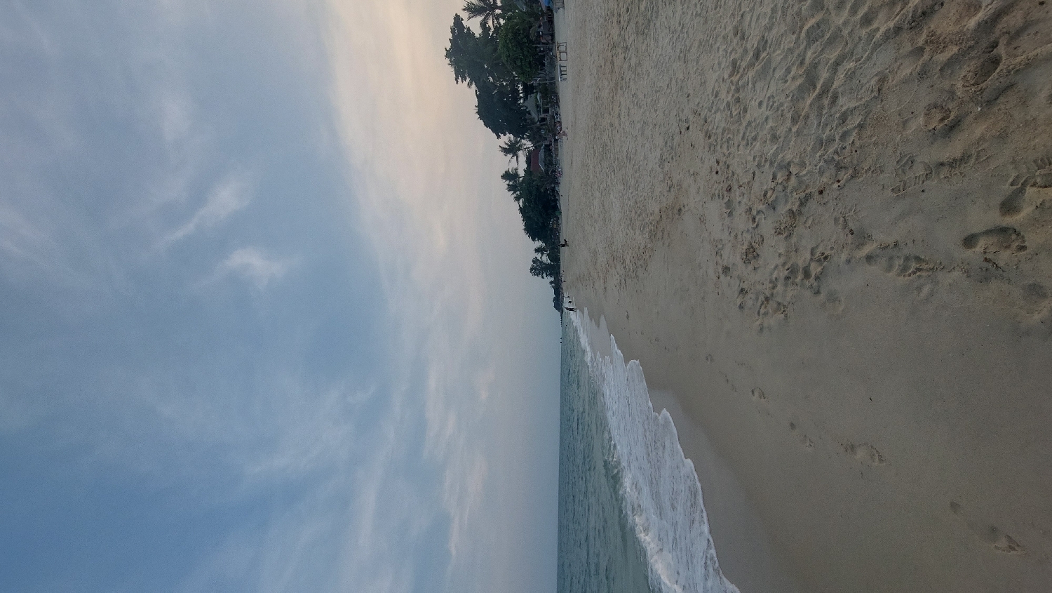 This media file presents a serene tropical beach scene, likely captured in Baan Lamai, Thailand, during either early morning or late afternoon. The image, which appears to be rotated 90 degrees clockwise, displays a wide expanse of light blue sky with soft, wispy white clouds diffused across it, hinting at a gentle, non-harsh light source.
The ocean occupies the middle ground, showcasing calm, blue-green waters with gentle white waves breaking rhythmically onto the shore. The foreground is dominated by a wide, light-colored sandy beach. Numerous footprints of varying sizes are visible in the sand, indicating recent human presence and activity along the shoreline.
Along the distant coastline on the left, a lush line of tropical vegetation, prominently featuring tall palm trees, stretches horizontally. Nestled among these trees, some indistinct structures or buildings can be seen, suggesting beachfront properties or local infrastructure. A few very small, distant figures are visible on the beach and in the water, enjoying the peaceful environment.
The overall mood is tranquil and natural, characteristic of a tropical beach setting. The soft lighting and the absence of harsh shadows suggest a time of day just after sunrise or before sunset. There is no visible text in the image.