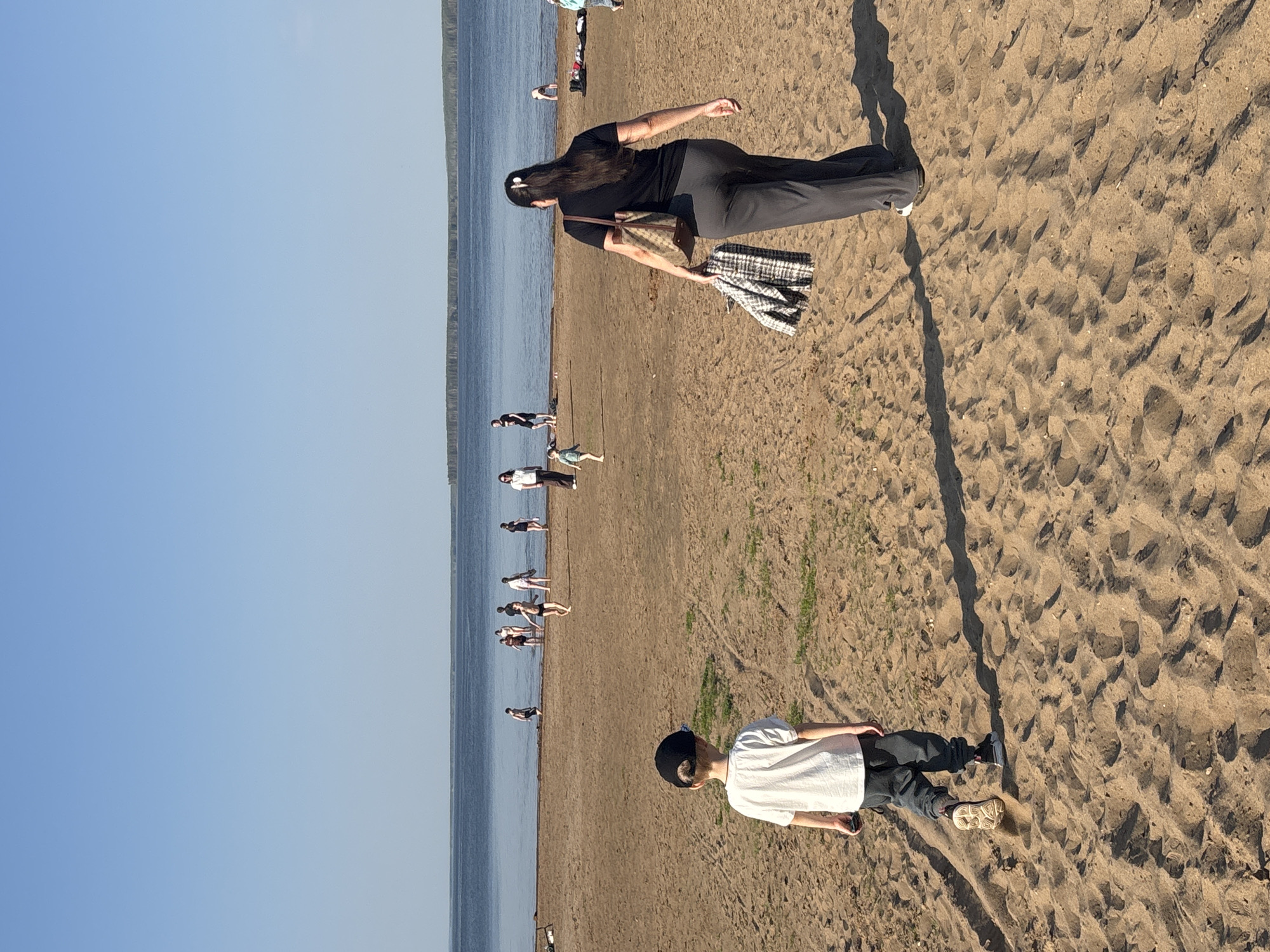 The image shows a beach scene in Chaikovsky, Russia. The sky is clear blue, meeting a body of water (likely the Kama River) at the horizon. Several people are walking and standing along the shoreline. In the foreground, a person wearing a white shirt, dark pants, a black beanie, and tan sneakers is standing on the sandy beach. Further up the beach, a woman wearing a black outfit, a handbag, and carrying a folded black-and-white fabric is walking. The sand texture is detailed, showing footprints and shadows. Some sparse vegetation is visible.